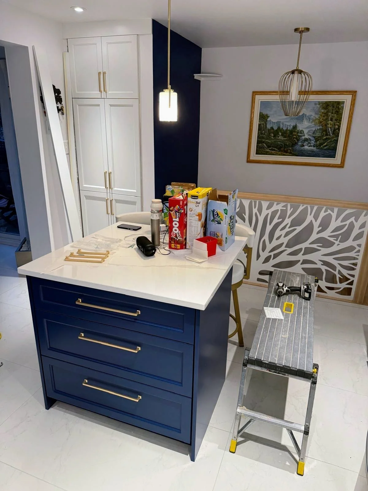 Kitchen island with blue base and white countertop, cluttered with cereal boxes, a water bottle, headphones, and other items. To the right, a step ladder with tools. In the background, white cabinets, a framed landscape painting, and a hanging pendan