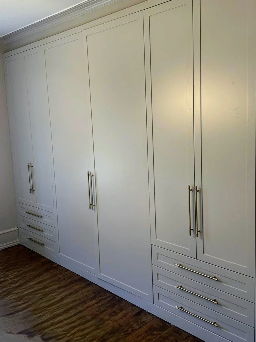 A large white built-in closet with multiple doors and drawers, featuring long metallic handles, situated against a wall with crown molding and a wooden floor.
