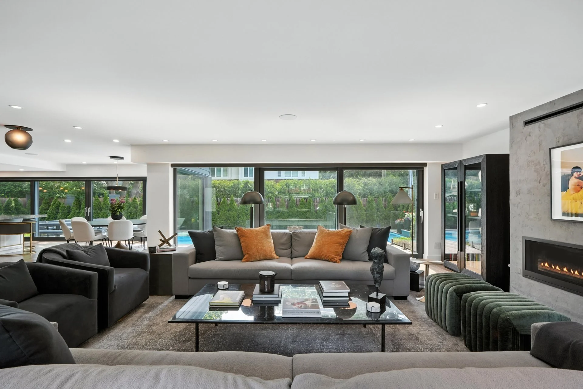 Modern living room with black and light gray sofas, orange throw pillows, glass top coffee table, fireplace, and large windows with greenery outside.