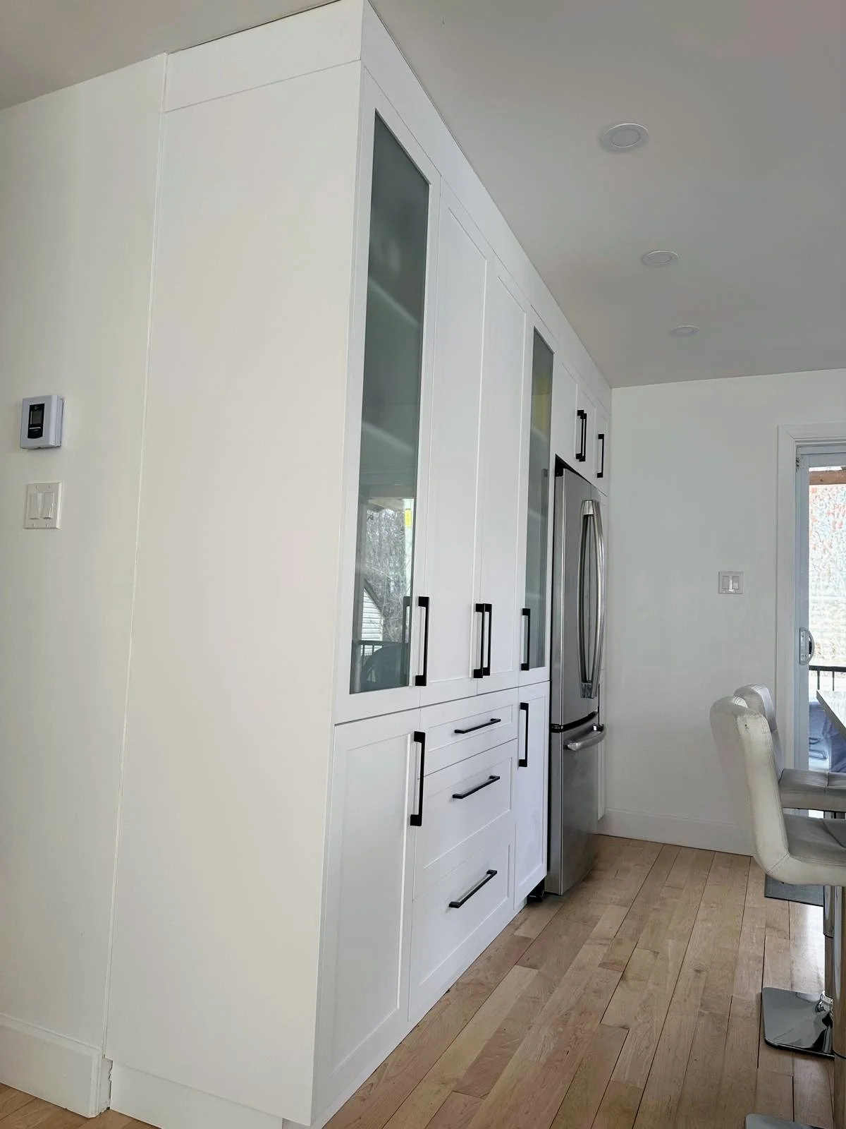 White kitchen cabinetry with black handles, stainless steel refrigerator, wooden floor, sliding glass door in the background.