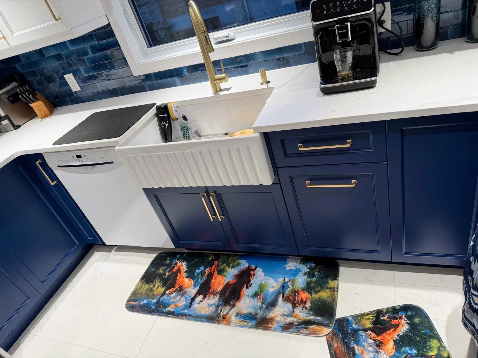 Blue kitchen cabinets with gold handles, white marble countertop, stainless steel sink with gold faucet, window above sink, black coffee machine with a glass cup, blue tiled backsplash, sofly colorful horse-themed rugs in front of cabinets and sink, 