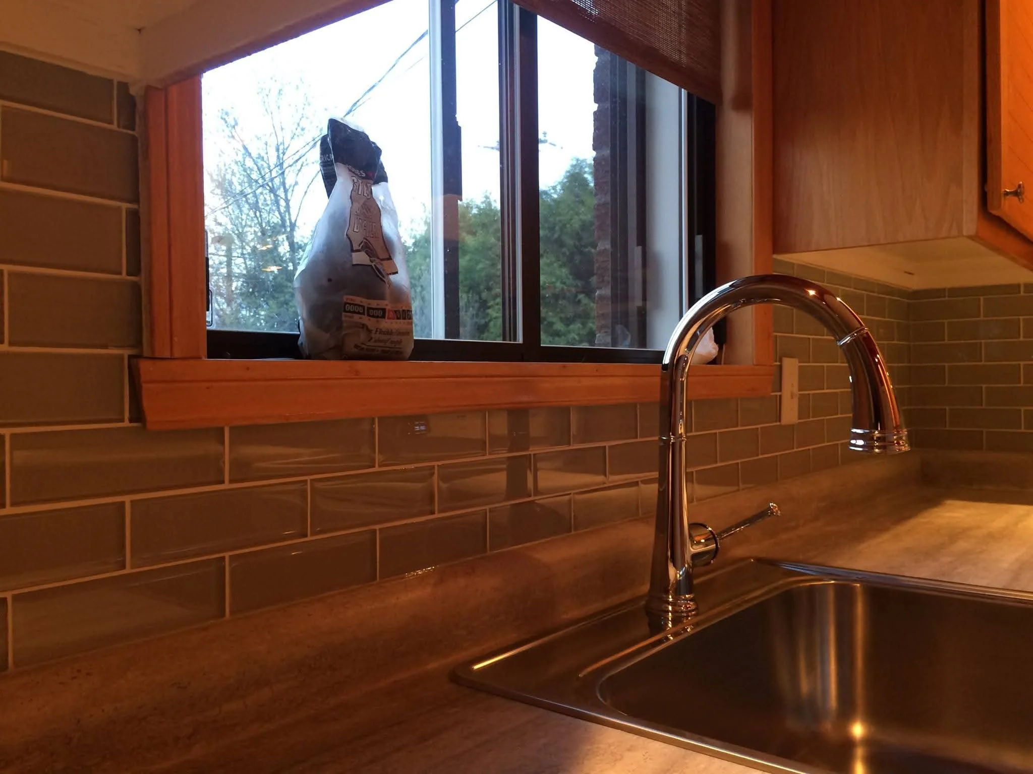 Kitchen sink with a shiny chrome faucet, beige backsplash tiles, and an open window with a paper bag outside, showing a cloudy outdoor scene with trees.