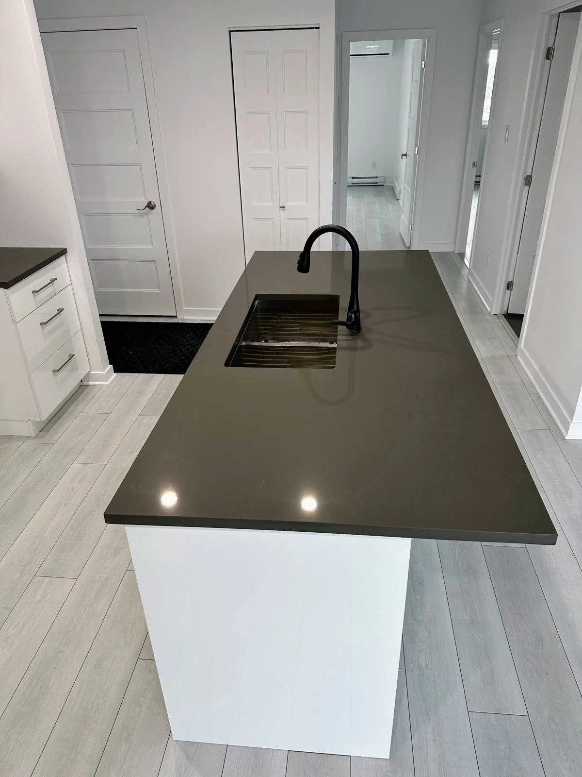 Modern kitchen island with a dark gray countertop and white base, black faucet, and built-in sink, with light-colored wood flooring and white walls.