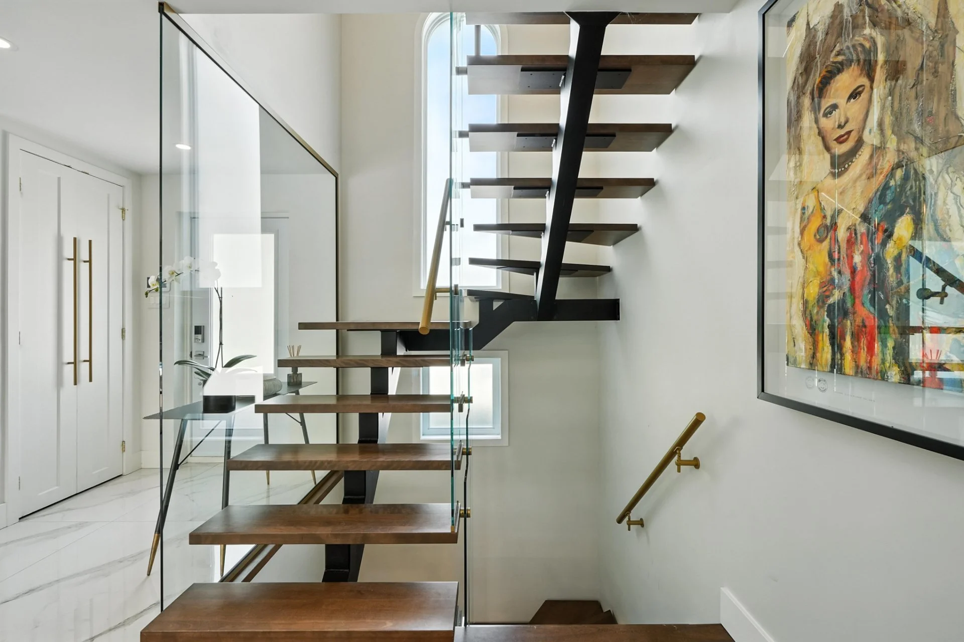 Modern interior staircase with wooden steps, black support, glass railing, and artwork on the wall, with white walls and natural light through window.