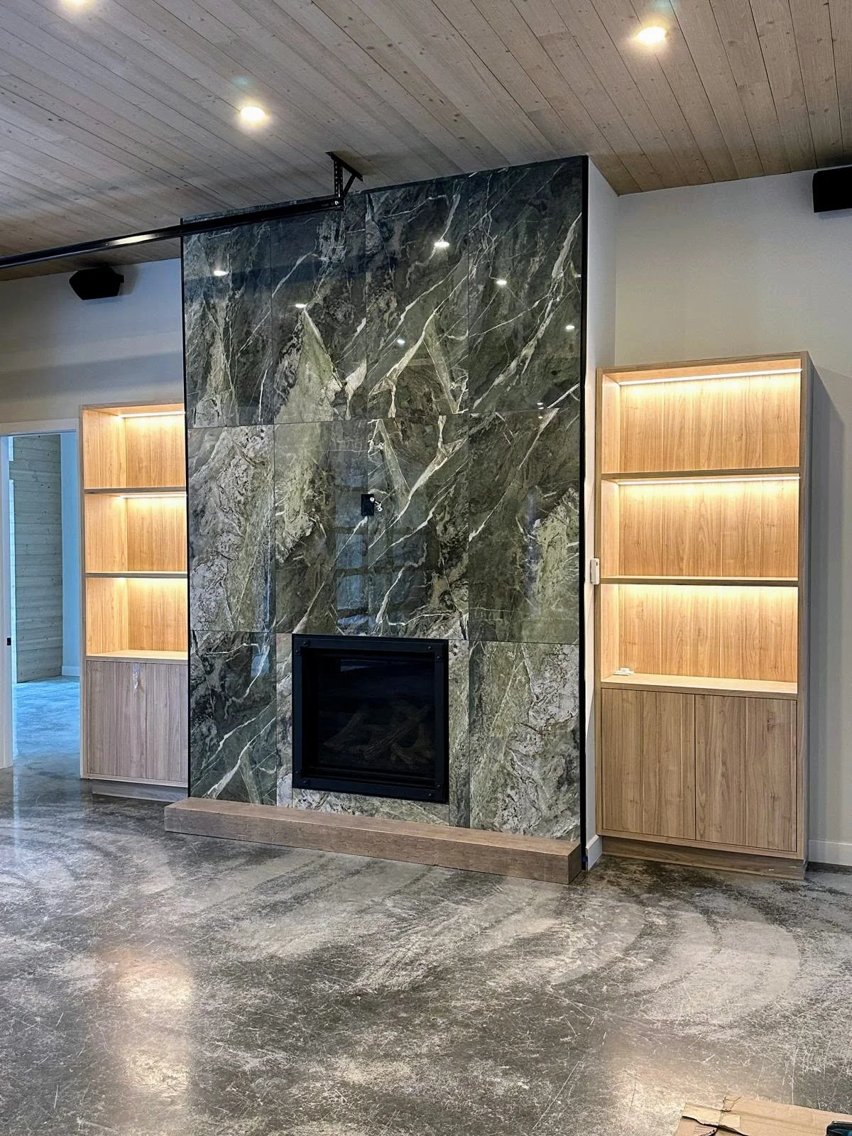 Living room features a dark green marble fireplace with a black glass door and two built-in wooden bookshelves with lighted shelves on either side, dark polished concrete floor, and a wooden ceiling with embedded ceiling lights.
