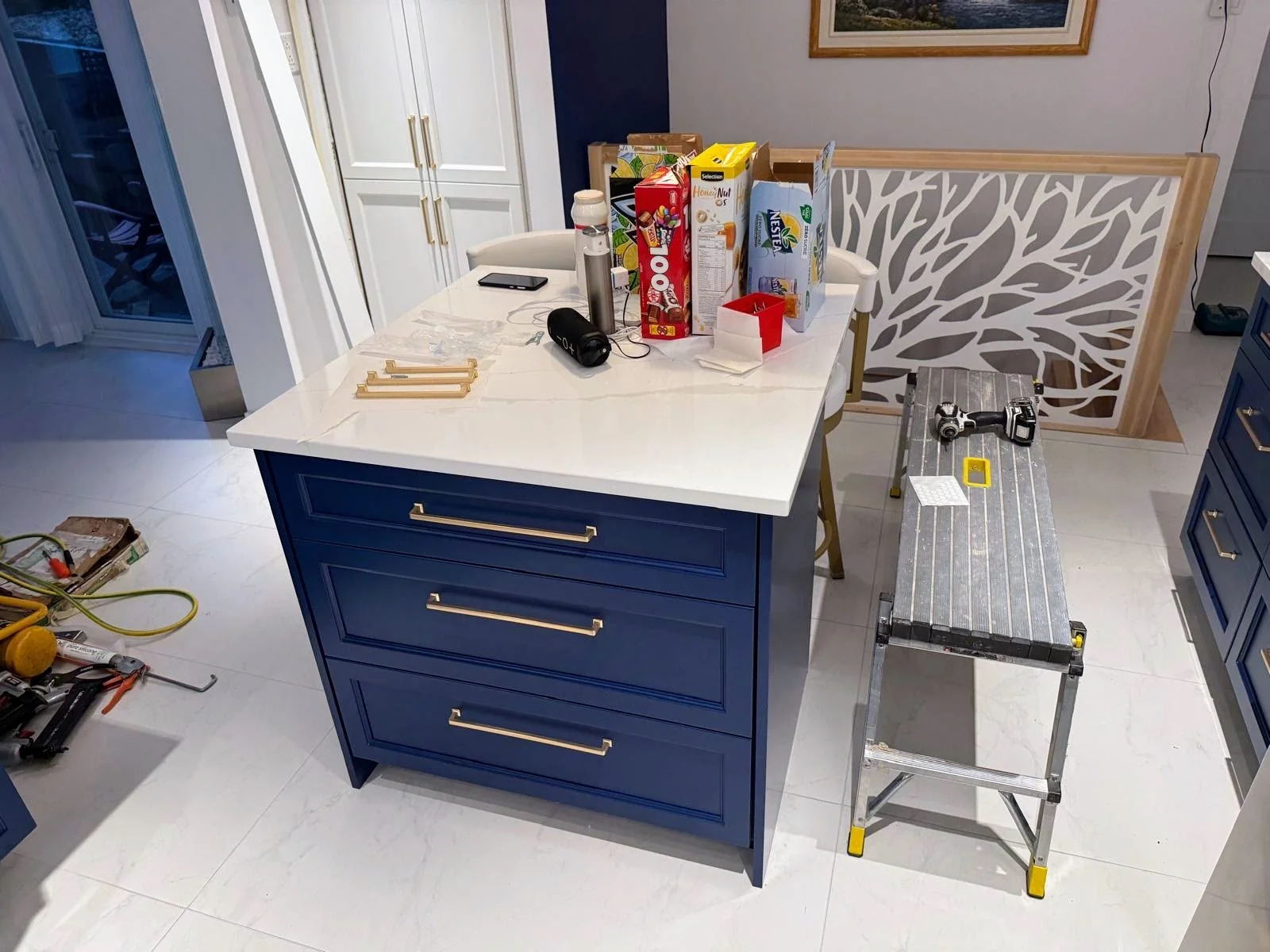 Kitchen island with a white marble top, blue drawers with gold handles, and a pack of various breakfast cereals on a white table. A handheld drill, measuring tape, and small white papers are on a silver step ladder. There are tools and yellow extensi