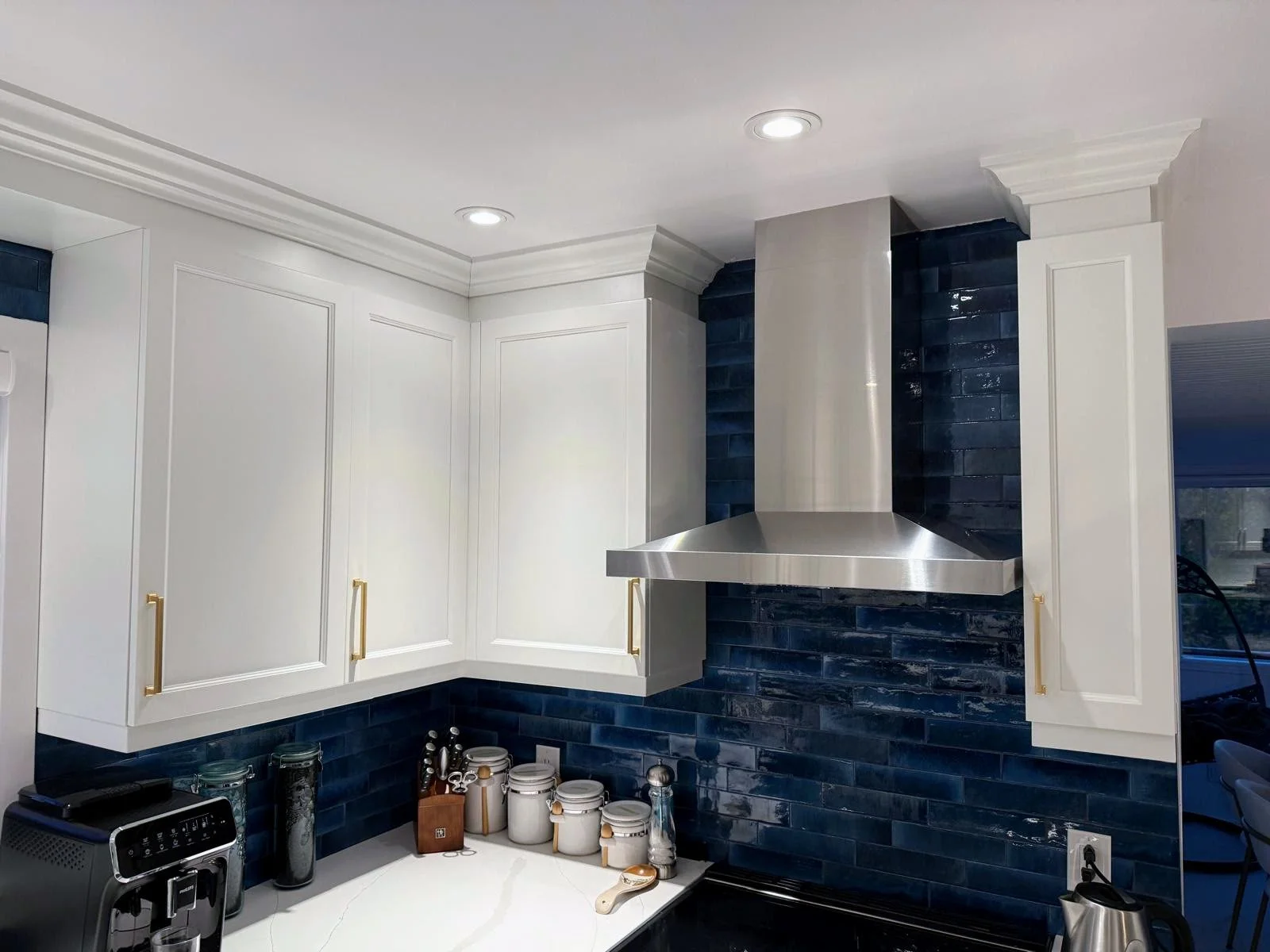 Kitchen with white cabinets, gold handles, dark blue tile backsplash, stainless steel range hood, and various kitchen appliances and containers on the counter.