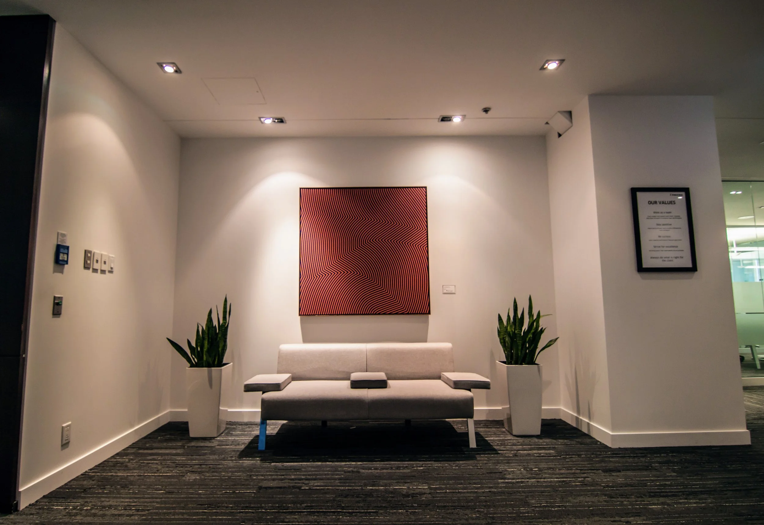 Modern office waiting area with a white sofa, two potted plants on either side, a red abstract art piece on the wall, and framed policy poster on the right wall.