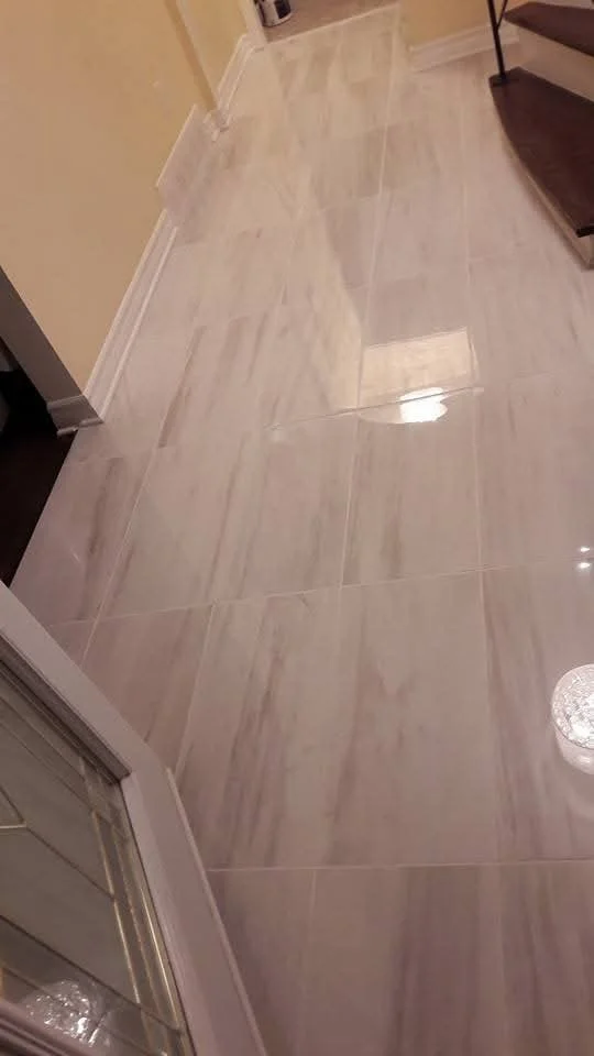 A hallway with beige walls, white baseboards, and a shiny beige tiled floor. A staircase with dark wooden stairs and metal railing is on the right. A small appliance or cabinet is visible in the background.