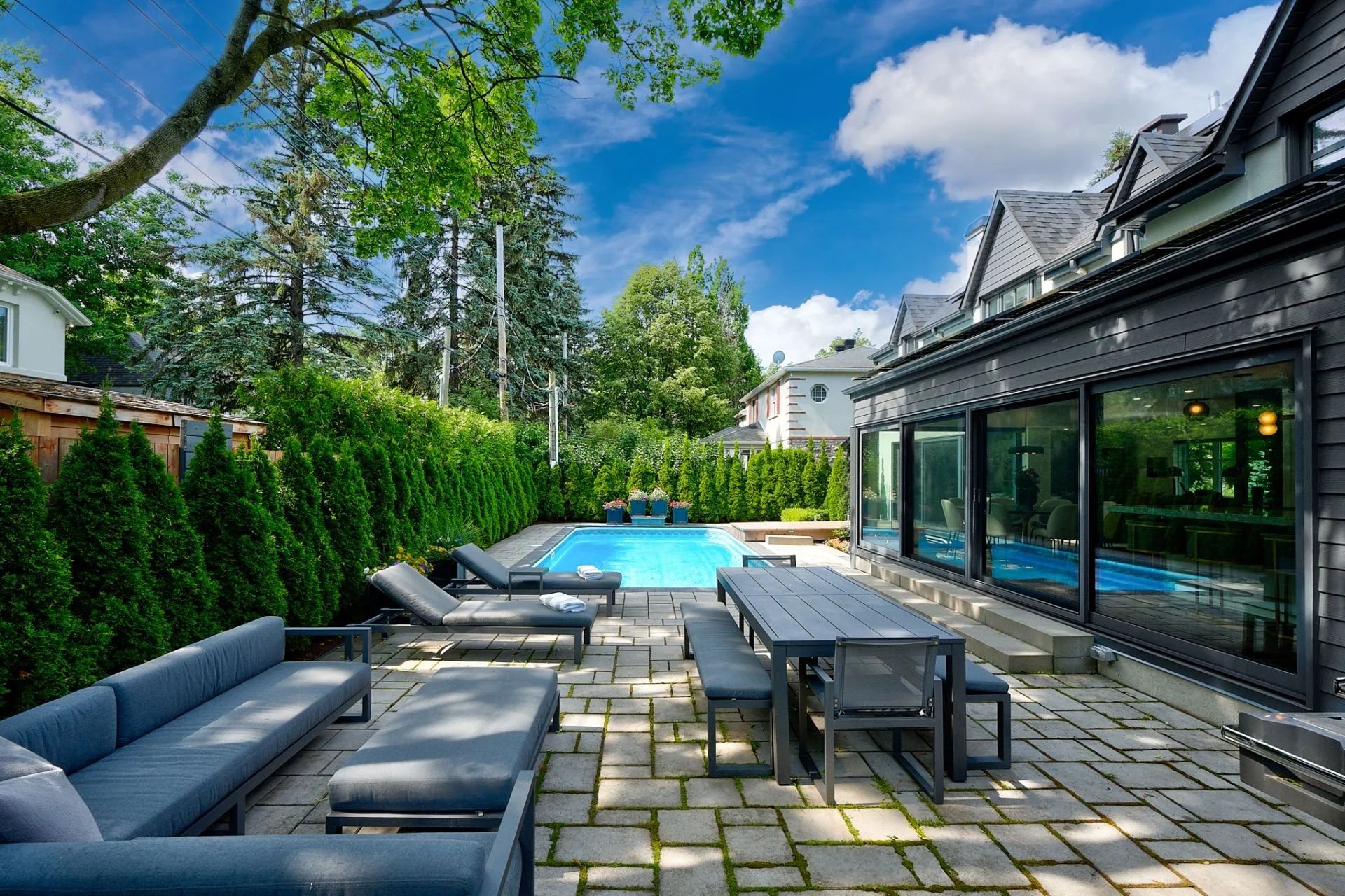 Backyard patio with outdoor furniture, a swimming pool, lush green trees and tall hedge in a suburban neighborhood on a clear sunny day.