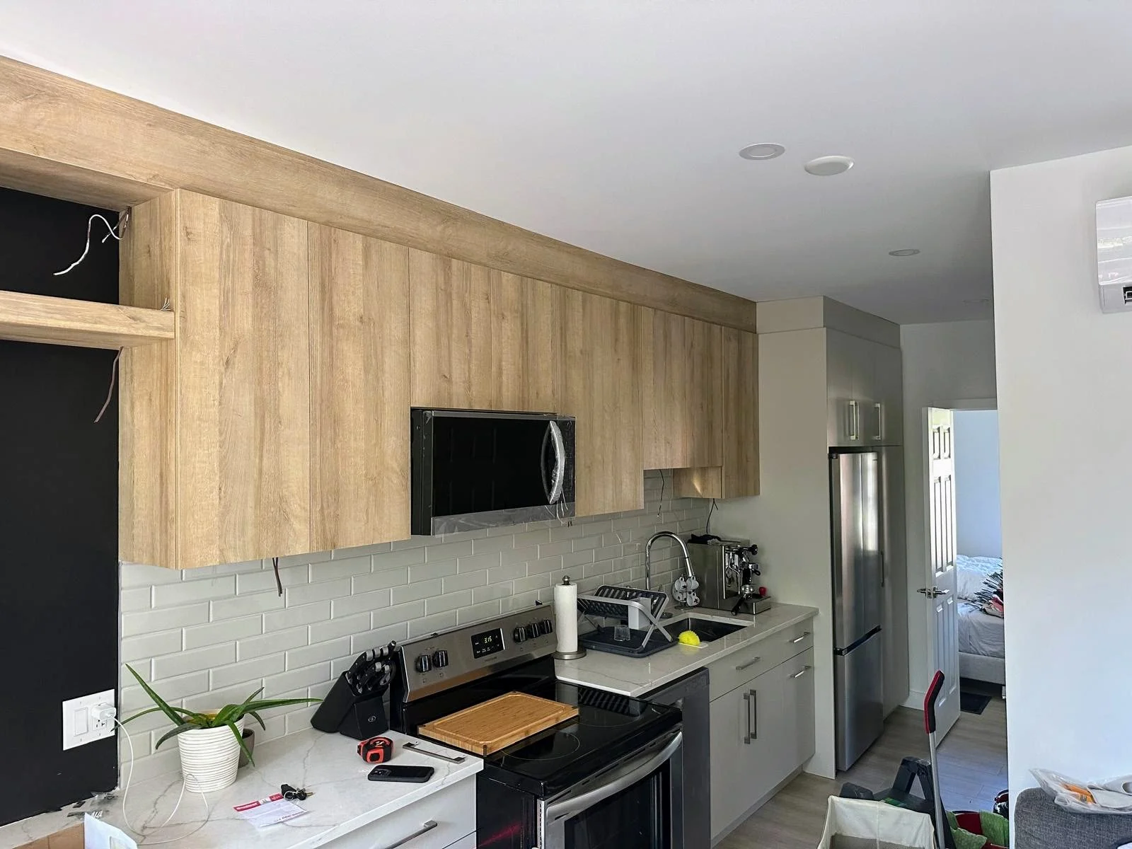 A modern kitchen with light wood upper cabinets, white lower cabinets, and grey subway tile backsplash. It has a stainless steel refrigerator, black stove, microwave, and various small appliances on the countertop. There is a houseplant, a cutting bo