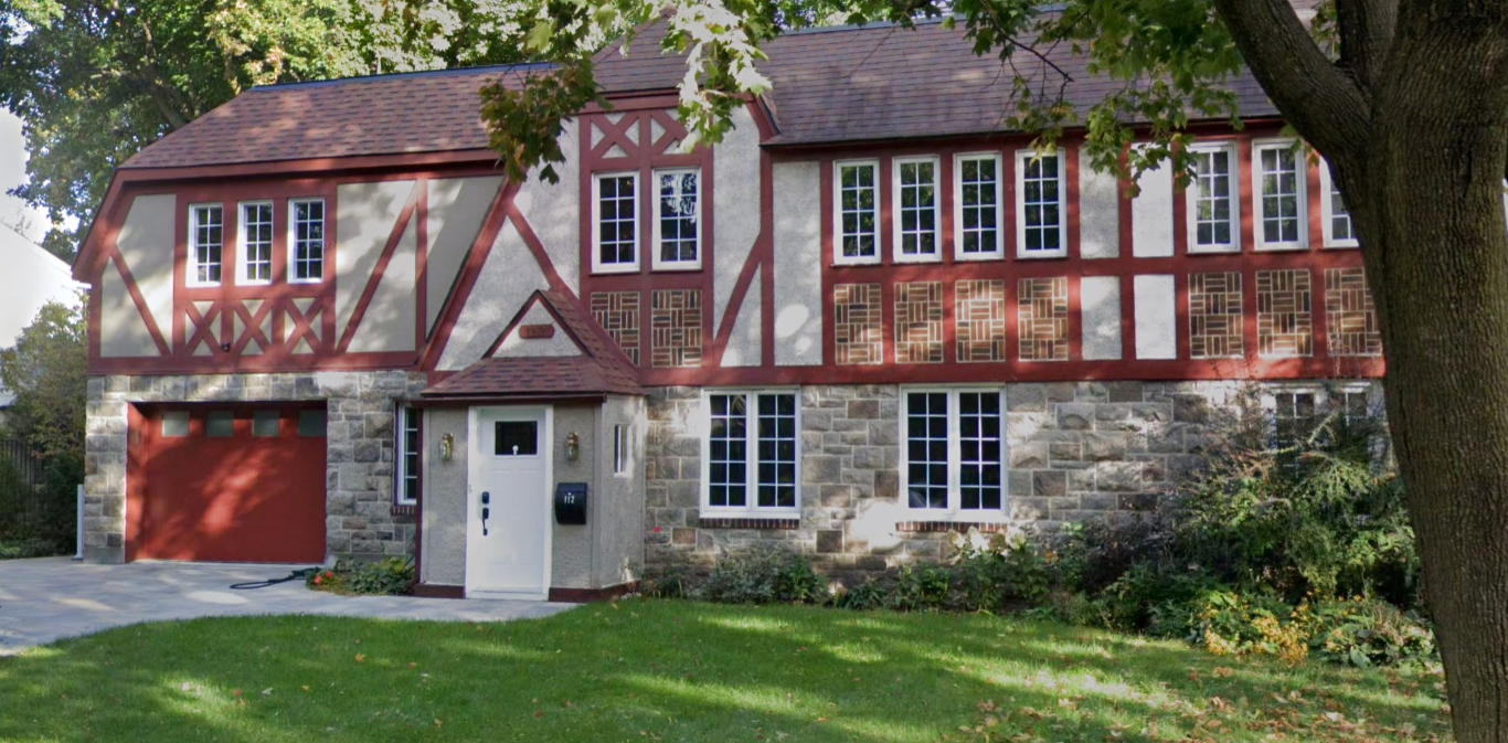 A two-story house with a stone foundation and half-timbered upper walls, a small front yard with grass, and a large tree partially obscuring the view.