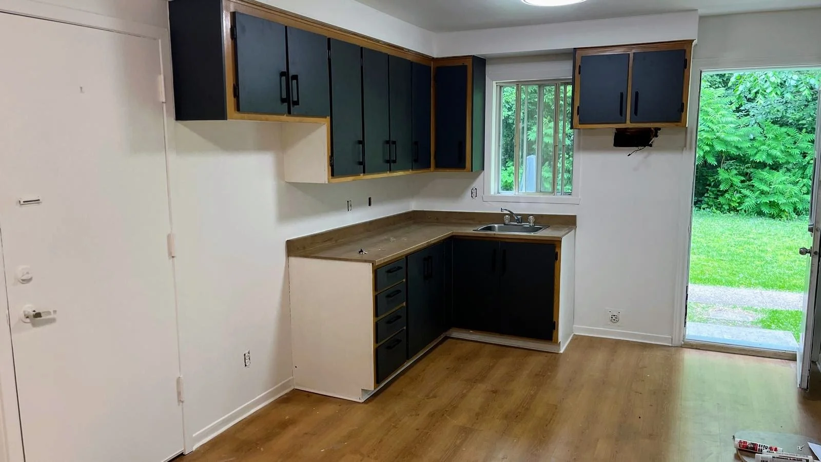 Empty kitchen with new black and wood cabinets, a countertop with a sink, window, and an open door showing a green lawn outside.
