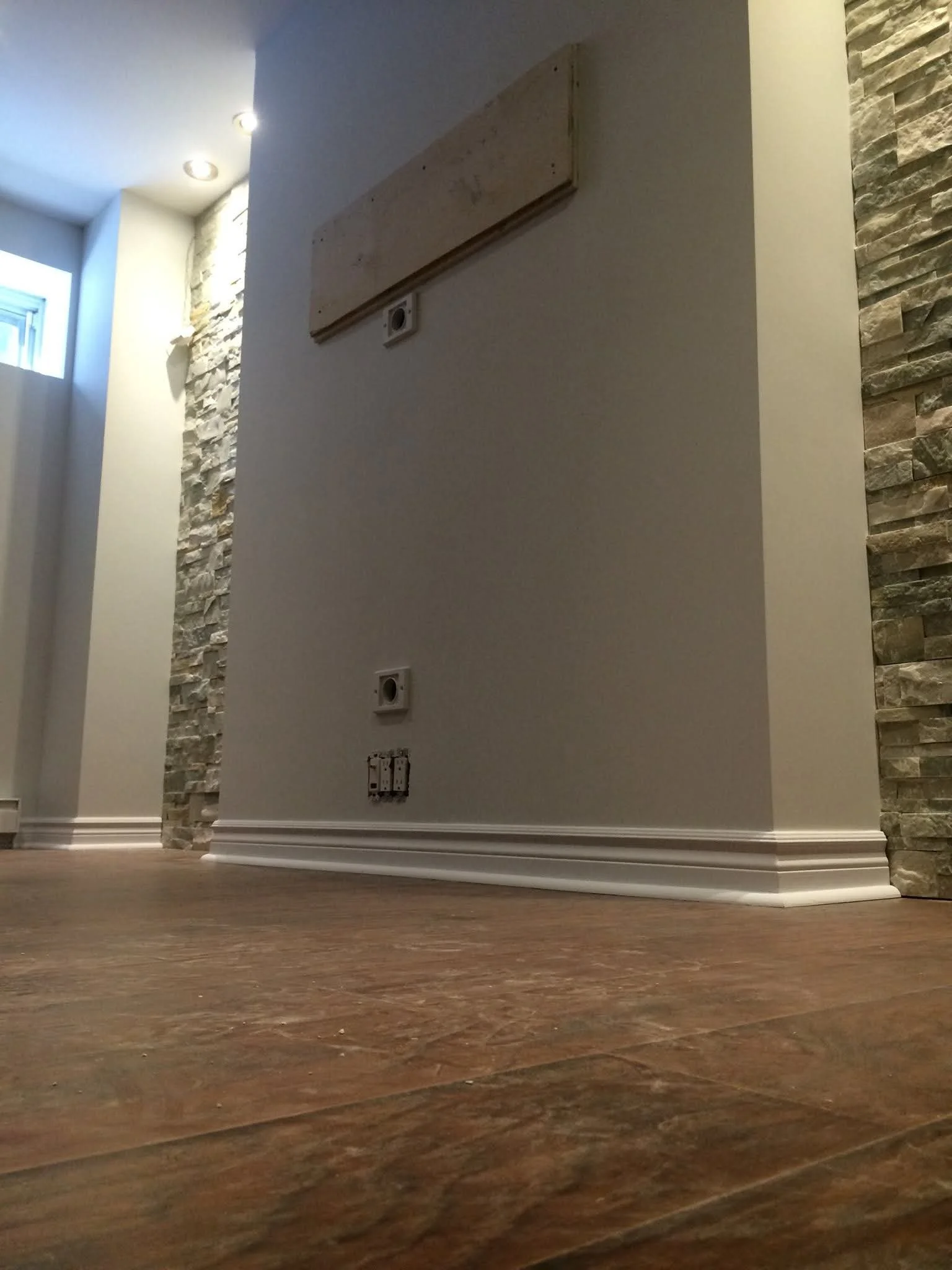 Interior wall with electrical outlets, a light-colored wooden board, and textured stone wall accents, with a window near the ceiling and recessed ceiling lights.