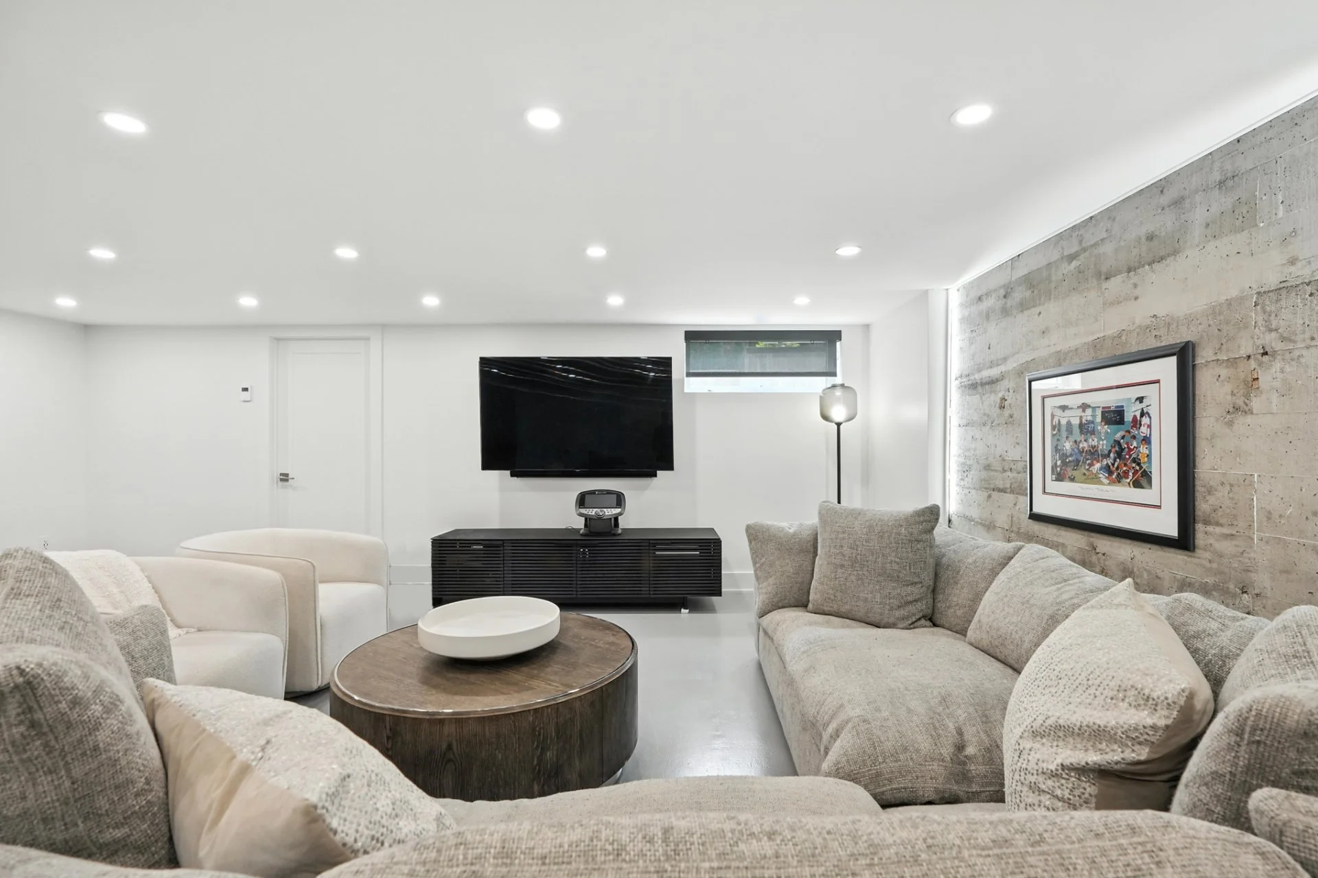 Modern living room with beige sofas, a wooden coffee table, a wall-mounted TV, a floor lamp, and a framed artwork on a textured concrete wall.
