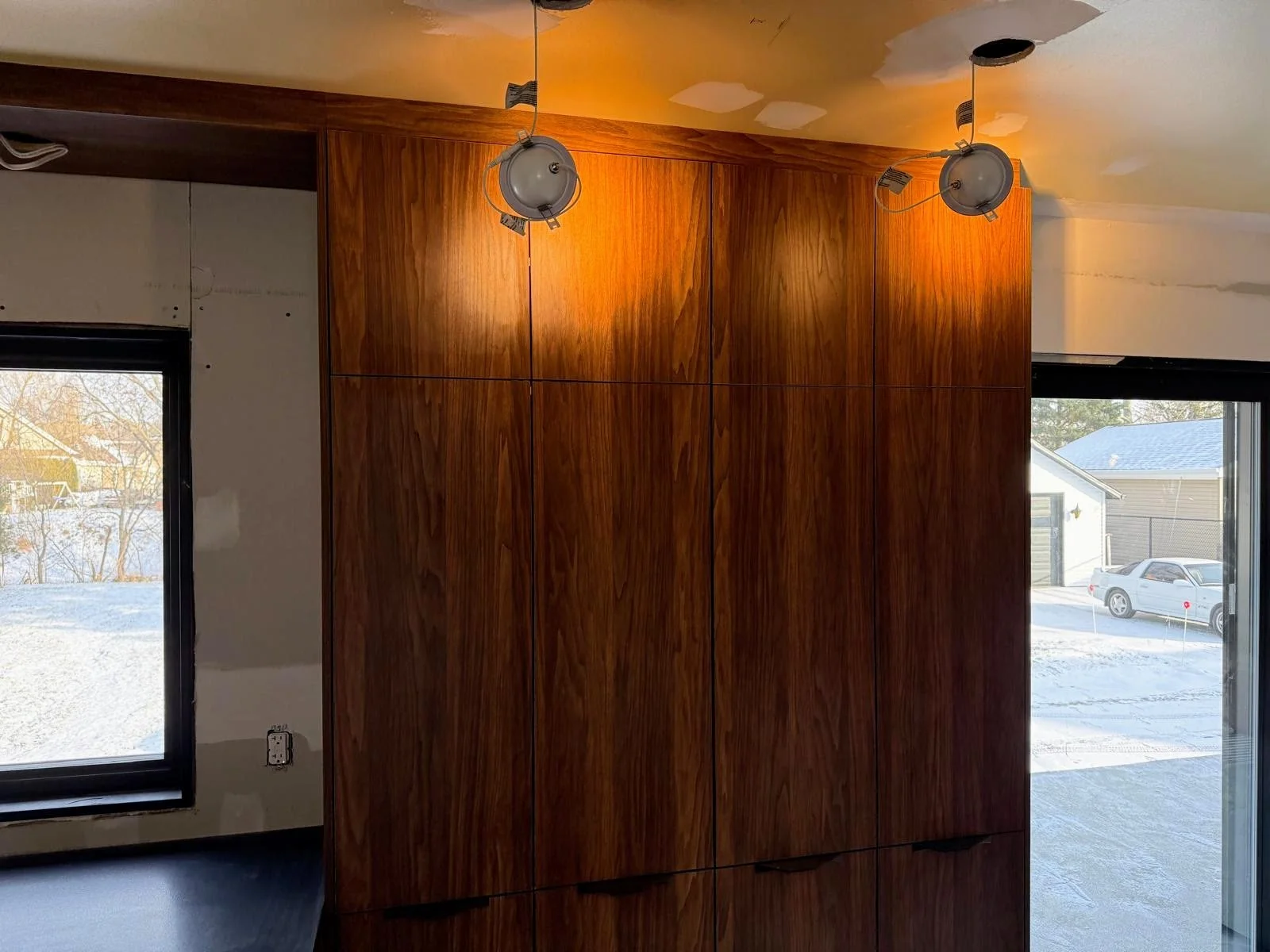 Interior of a room with a large wooden cabinet, two ceiling lights, a window showing a snowy outdoor scene, and a sliding glass door.
