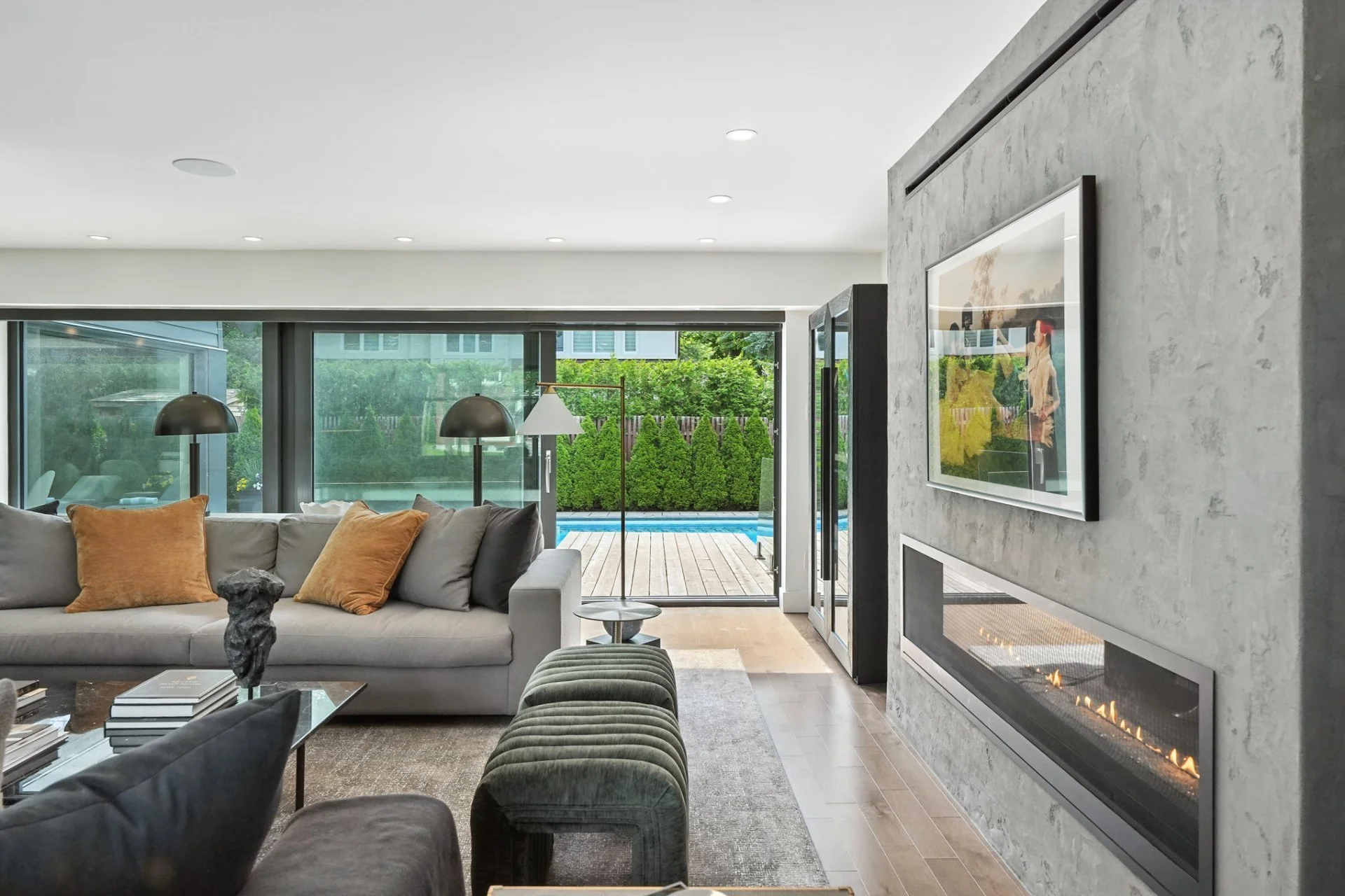 Modern living room with grey sofa, brown and grey pillows, glass coffee table, and a fireplace installed in a textured grey wall. Large sliding glass doors lead to a backyard with a pool and green foliage.