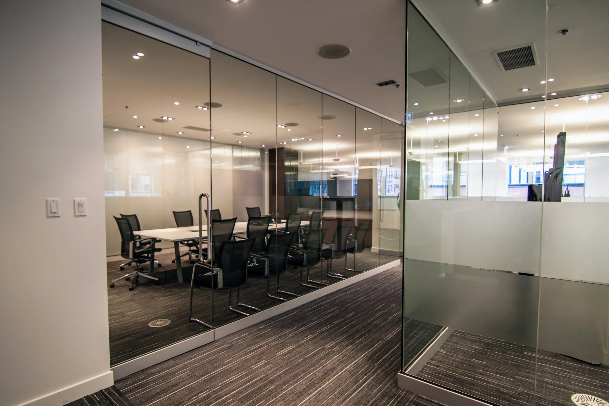 A modern conference room with a rectangular white table surrounded by black office chairs, enclosed by glass walls and doors, with ceiling lights and a view of a bright office space outside.