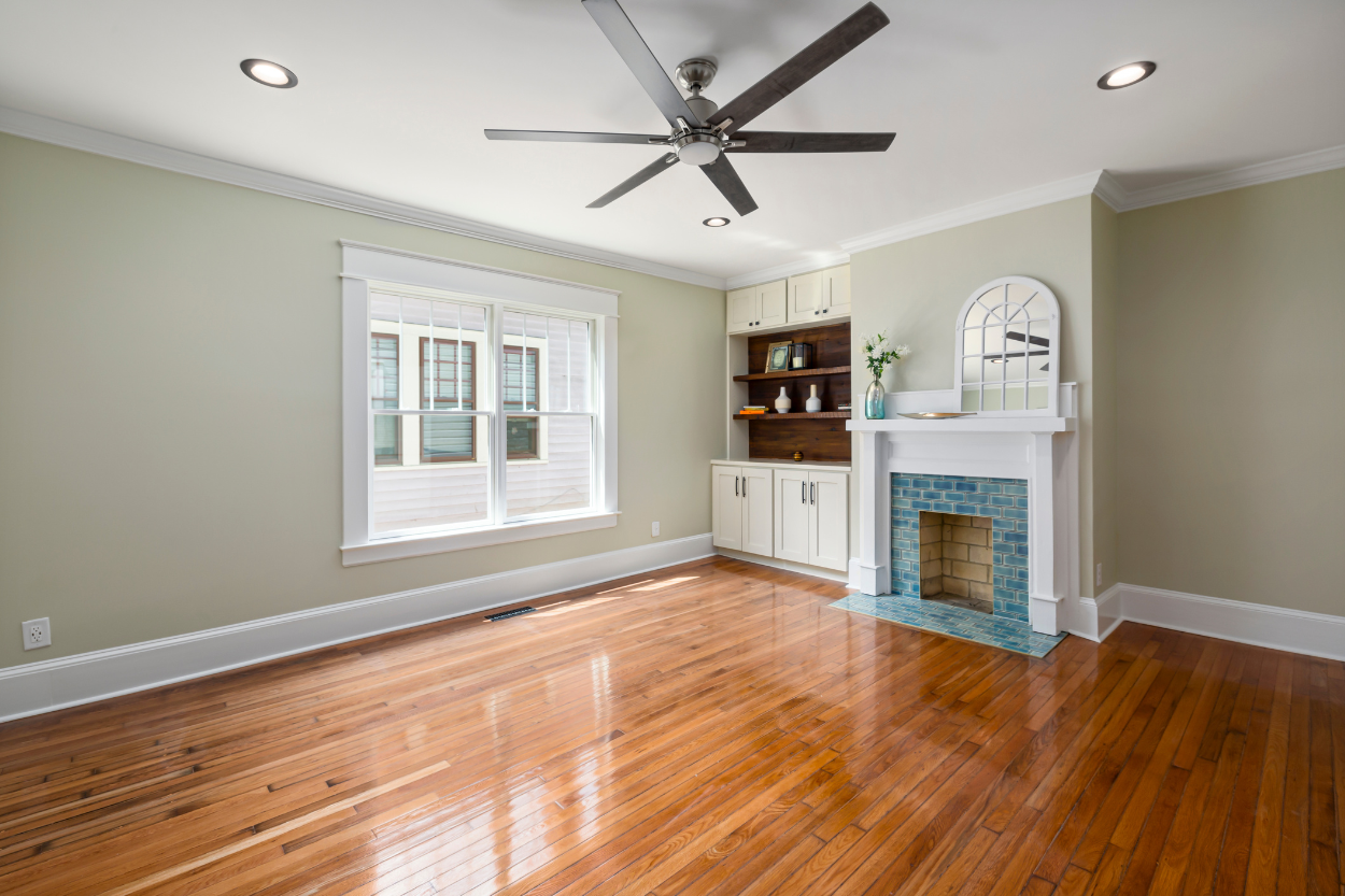 Empty living room with hardwood floors, a window, and a fireplace with a white mantel and blue tile inside.