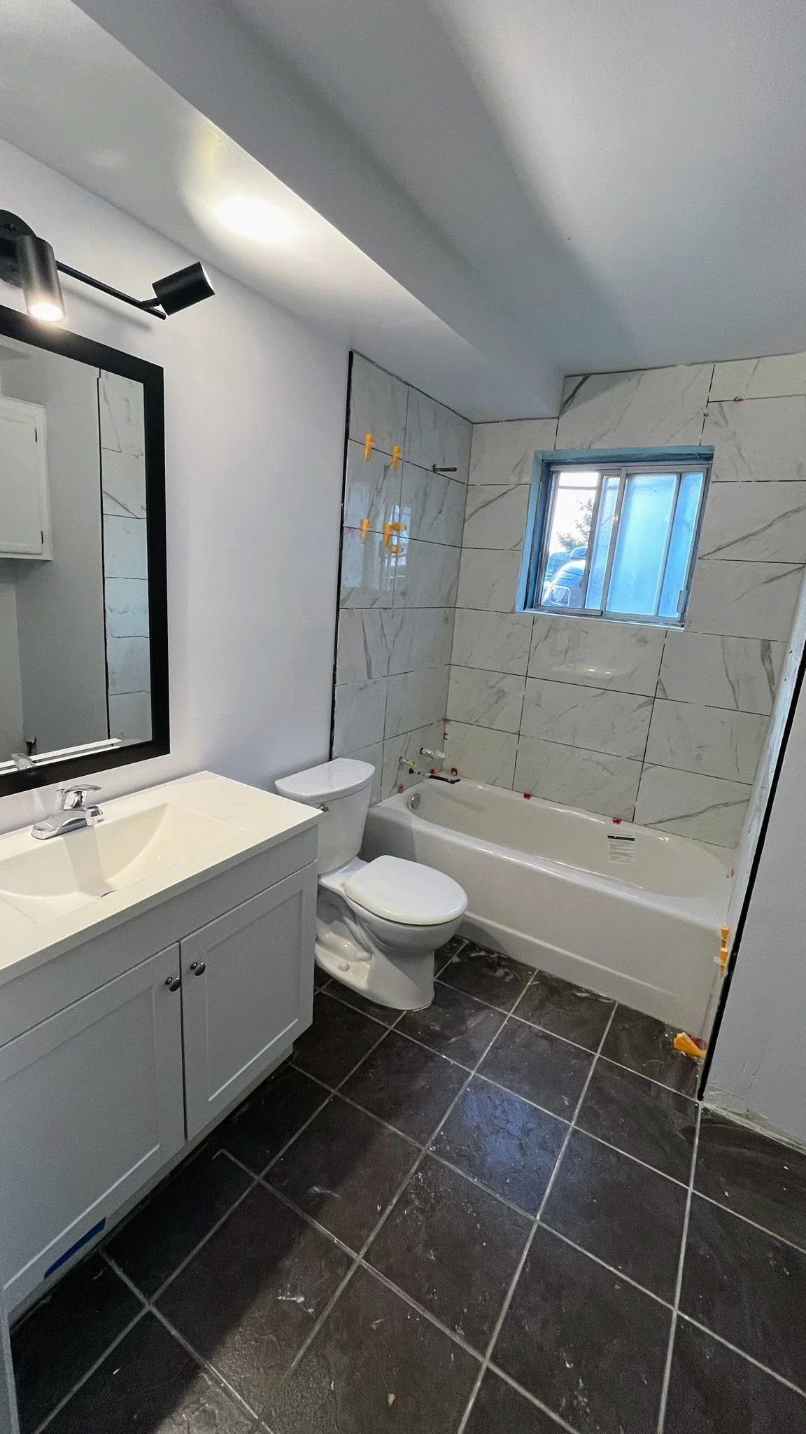 Bathroom undergoing renovation with white vanity, black-framed mirror, toilet, bathtub, and a small window, with construction markers on tiles and a dark tiled floor.