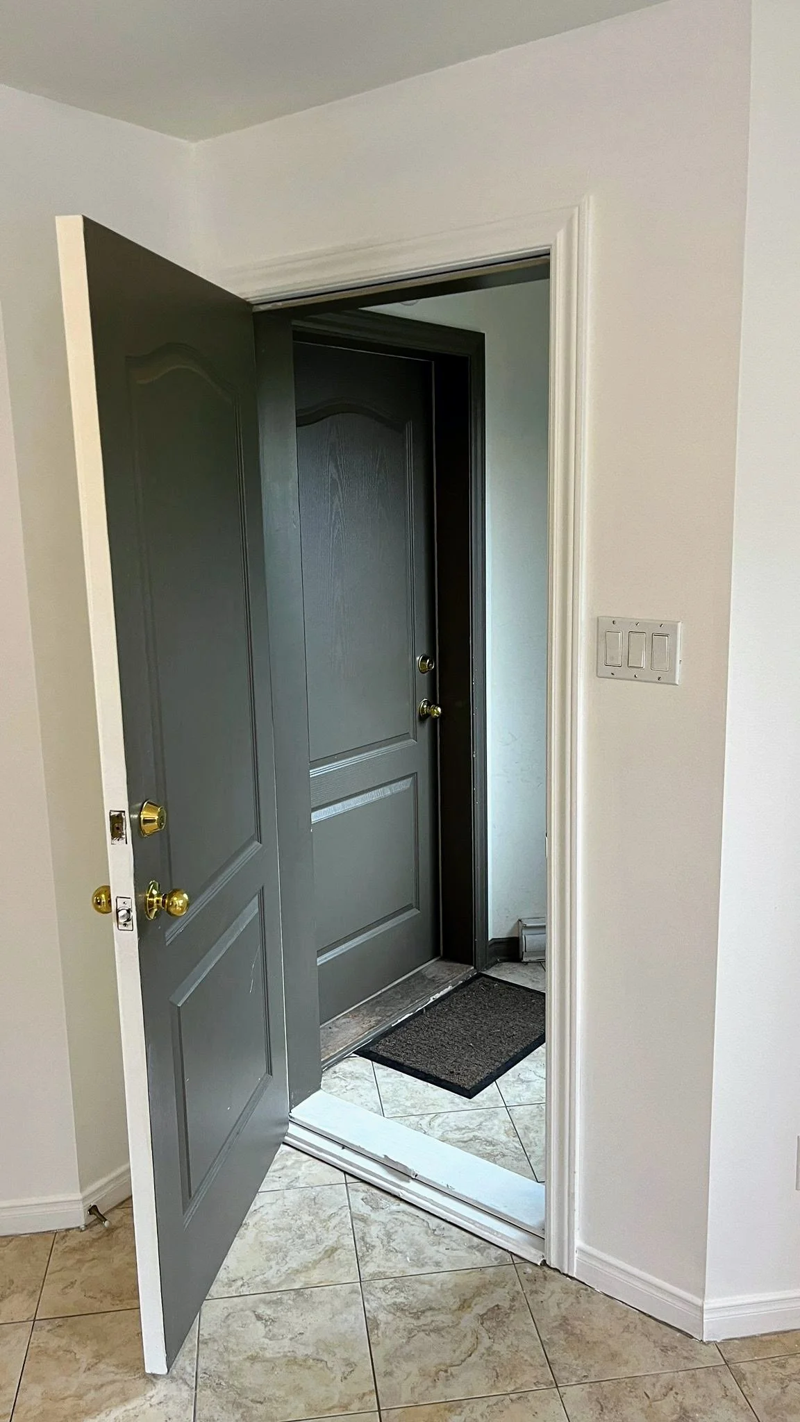 Open front door revealing black door with gold doorknob, leading outside or to another room. Gray tiled floor inside, with black doormat at threshold. White walls and trim, light switch on right wall.