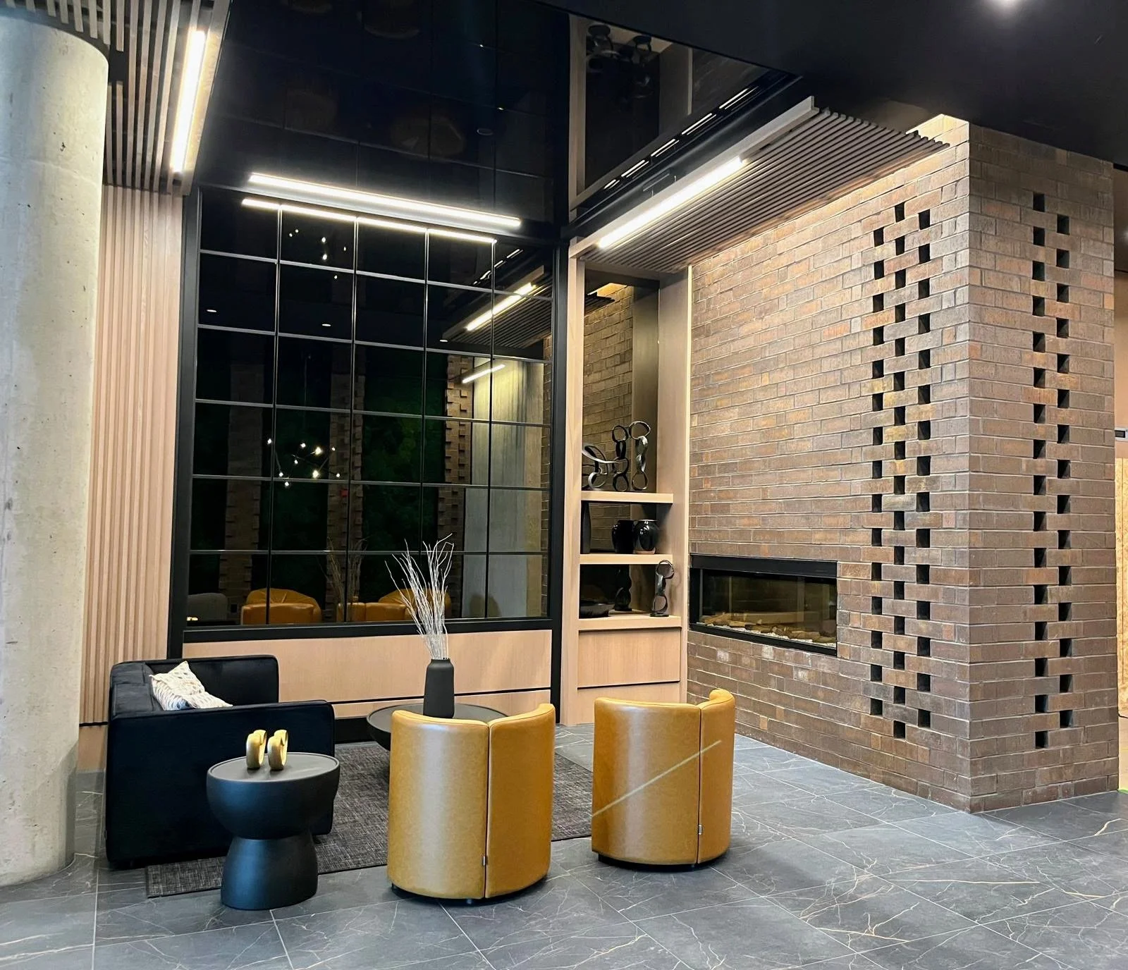 Modern hotel lobby area with black sofa, yellow chairs, a small round black side table, a decorative vase with white branches, a built-in fireplace, shelves with decorative objects, a large window with grid pattern, and contemporary design elements.