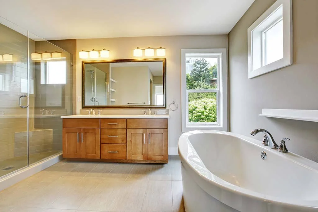 A modern bathroom with a glass-enclosed shower, a large mirror above a wooden vanity with four drawers, a bathtub near two windows, and neutral-colored walls and floors.