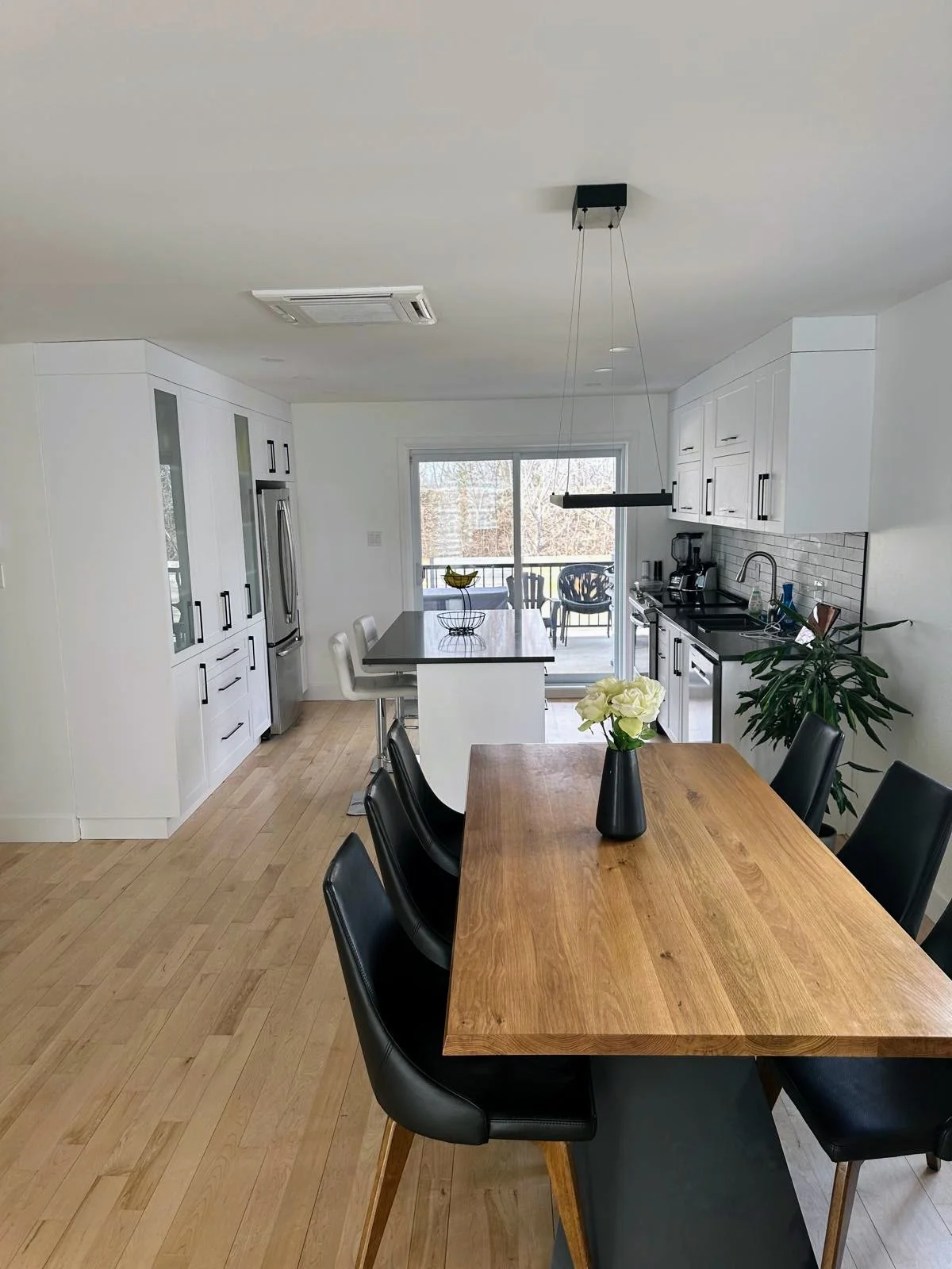 Modern kitchen and dining area with white cabinetry, a wooden dining table with six black chairs, an island with bar stools, and sliding glass door leading to a balcony.