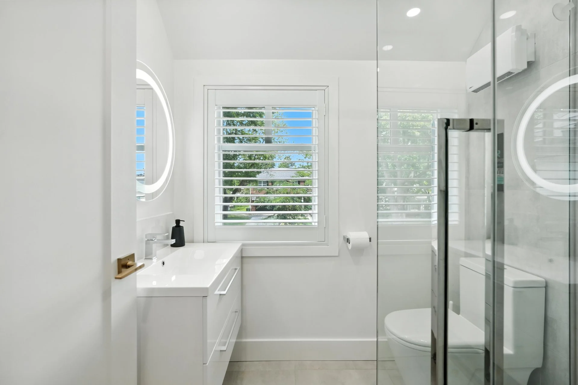 Modern white bathroom with a window, mirror, vanity, toilet, and shower enclosure with glass door.