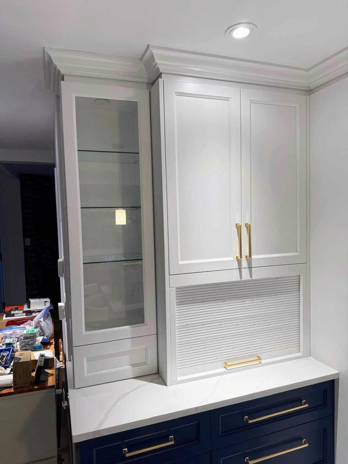 A kitchen cabinet with white upper cabinets and a blue lower cabinet, featuring gold handles, and a marble countertop. The upper cabinets include a glass front display section and a shaded roll-up door.