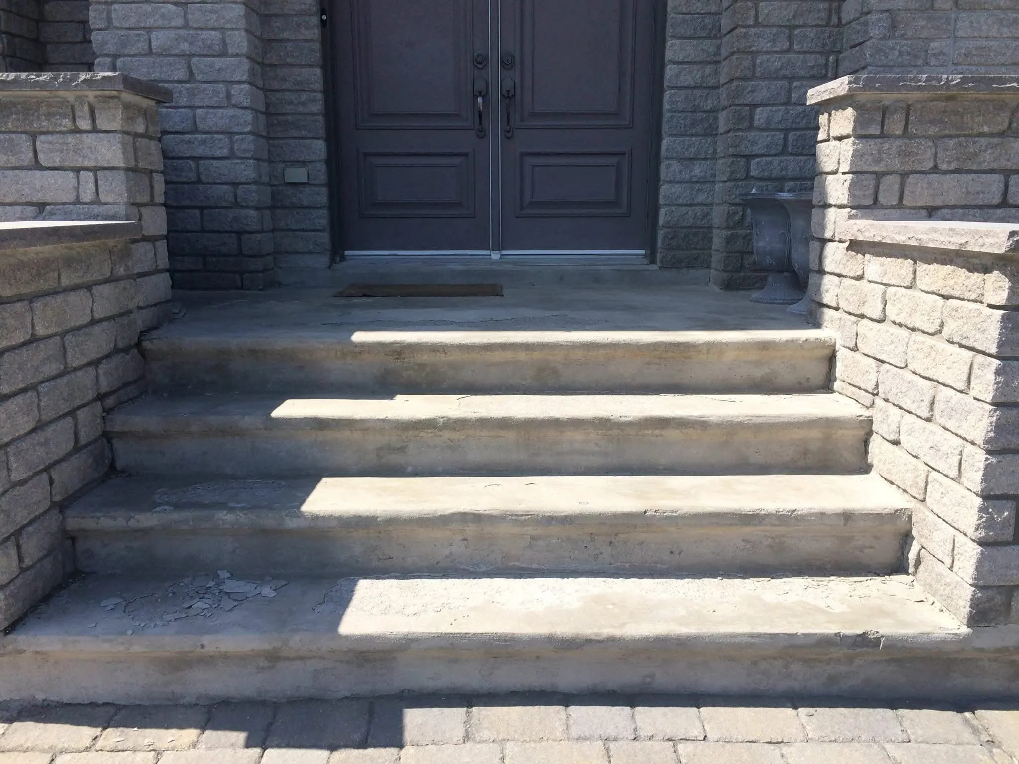 Concrete stairs leading up to a dark front door, with brick walls on either side and a small doormat in front of the door.