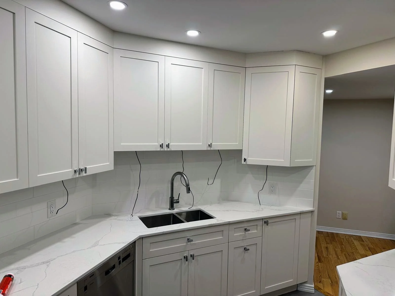 White kitchen cabinets with silver handles, black double sink, white marble countertops, and hardwood floors, with recessed ceiling lights.