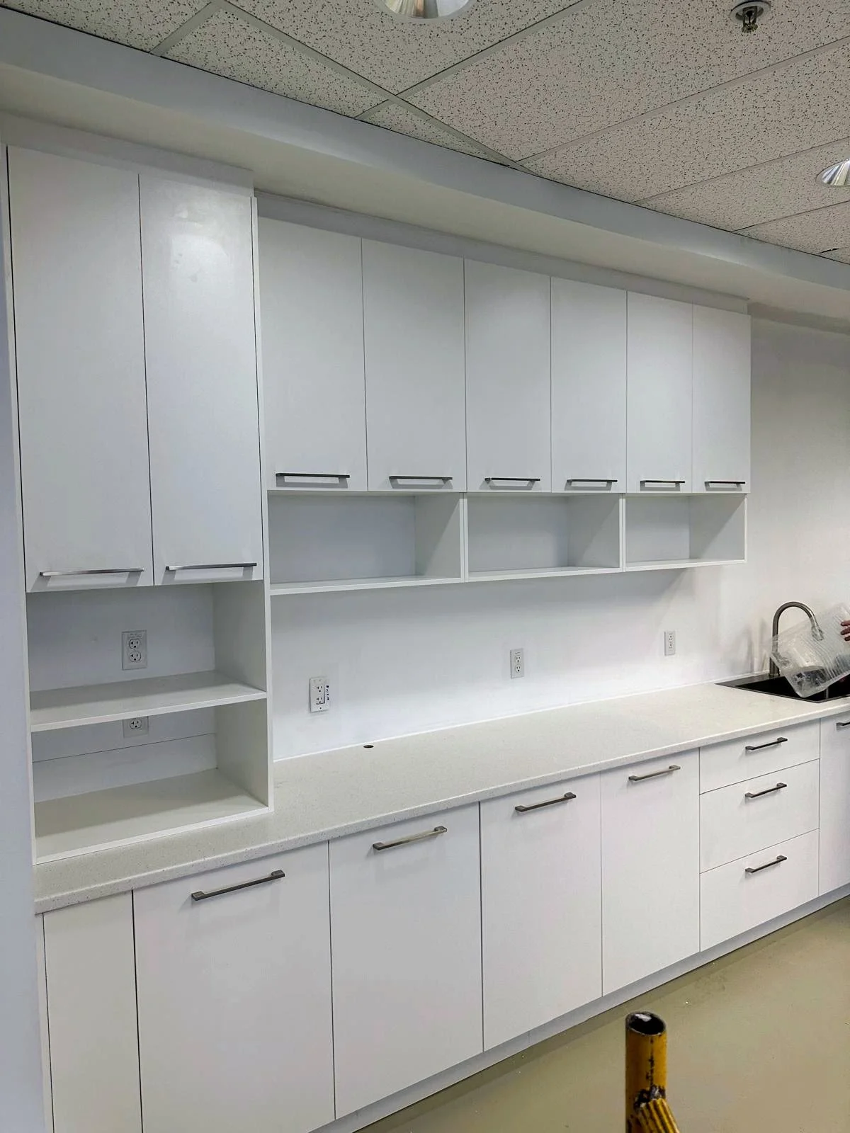 Empty white kitchen cabinets and countertop with a sink on the right, a person handling a plastic container by the sink, and multiple electrical outlets above the countertop.