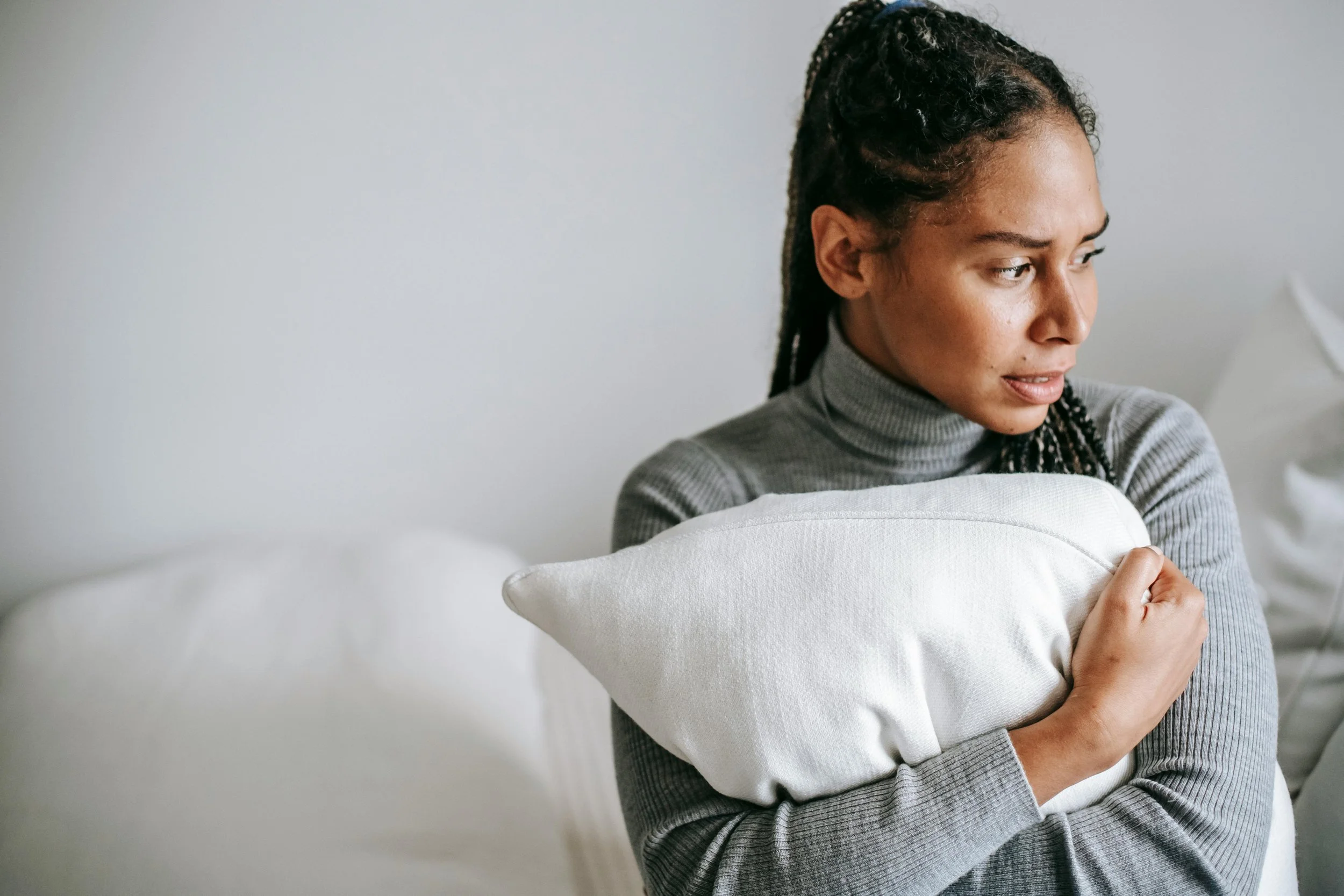 A woman with braided hair wearing a gray turtleneck sweater holding a white pillow close to her chest, looking to her right with a contemplative expression.