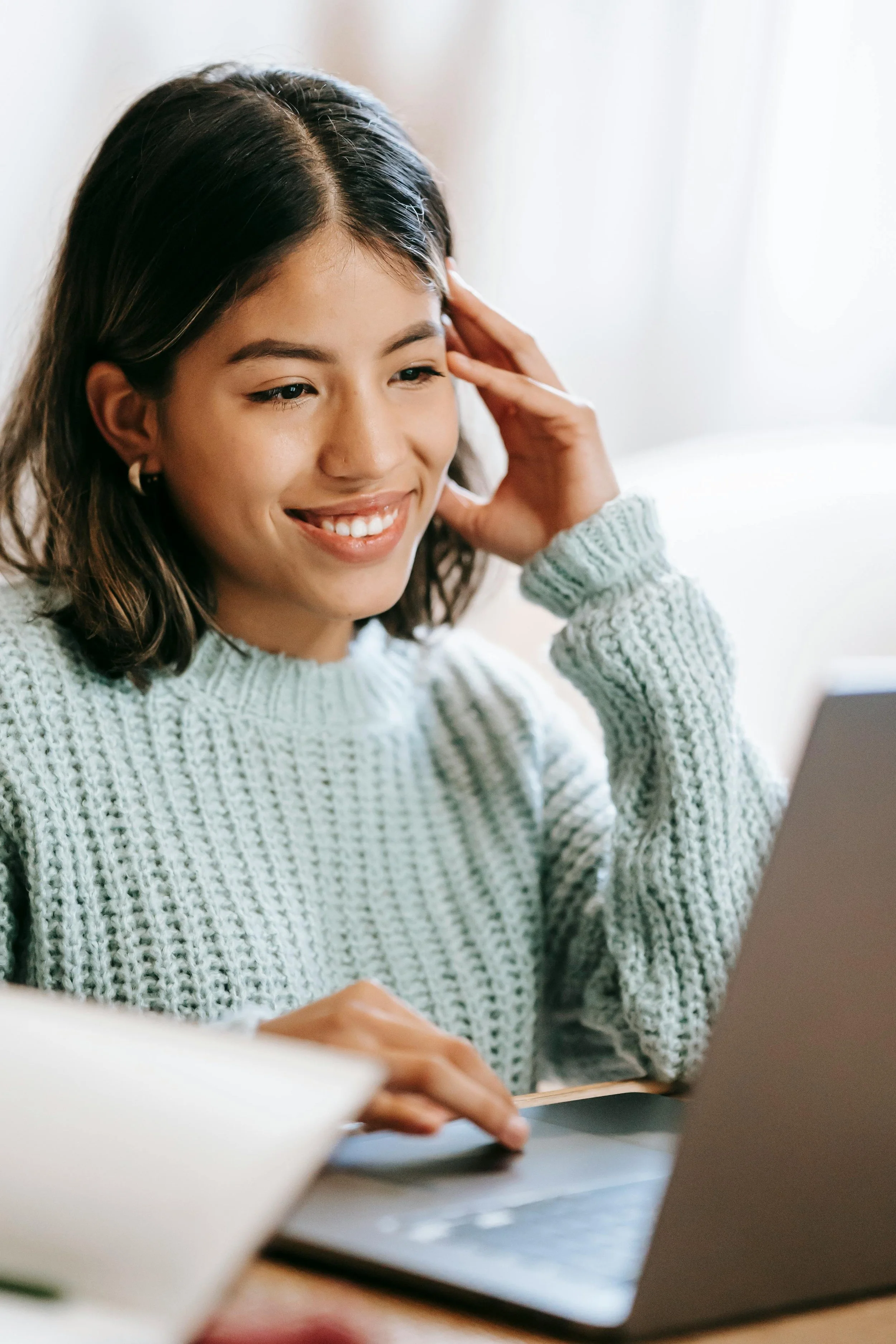 A young woman with shoulder-length brown hair, wearing a gray knitted sweater, smiling and looking at her laptop in a well-lit room.