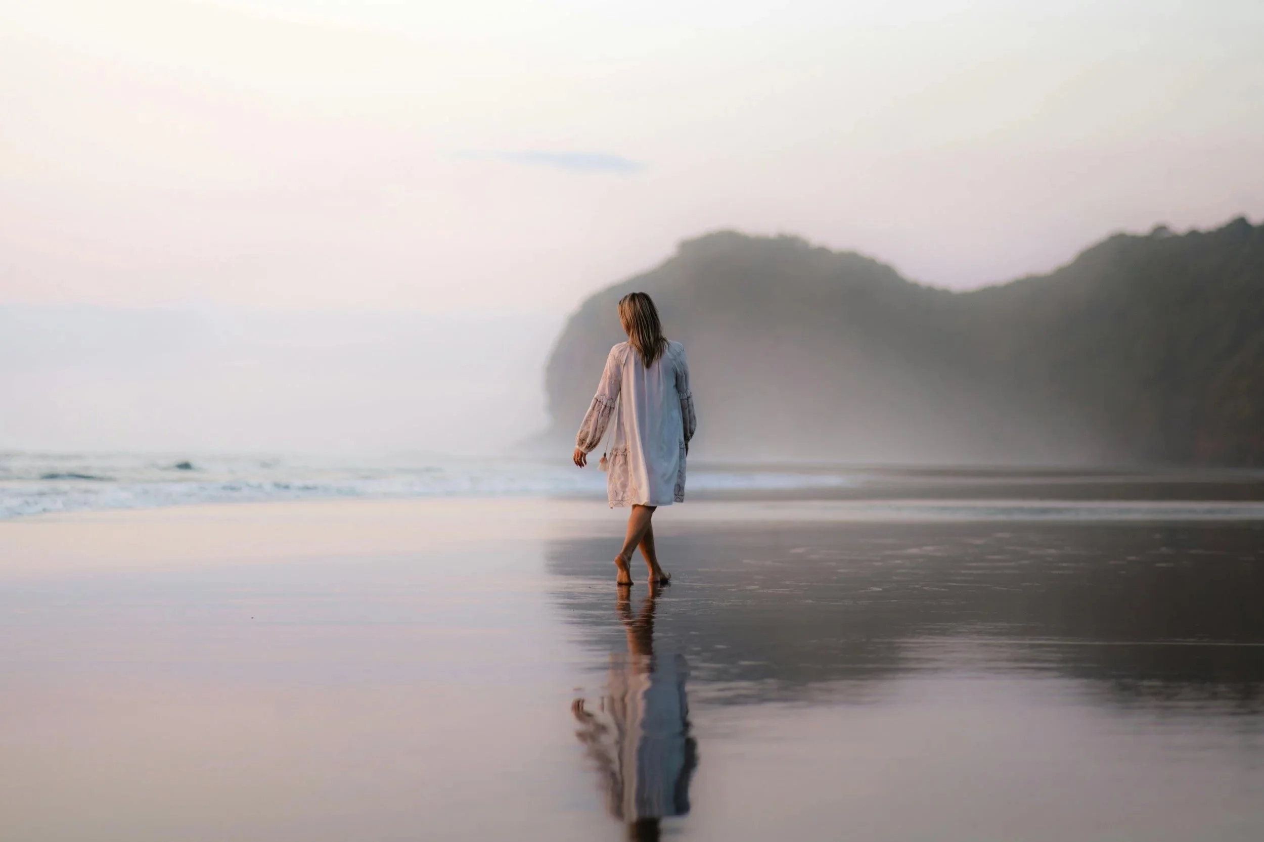 A woman in a white dress walking barefoot on a wet beach during sunset, with large cliffs in the distance and her reflection visible on the sand.