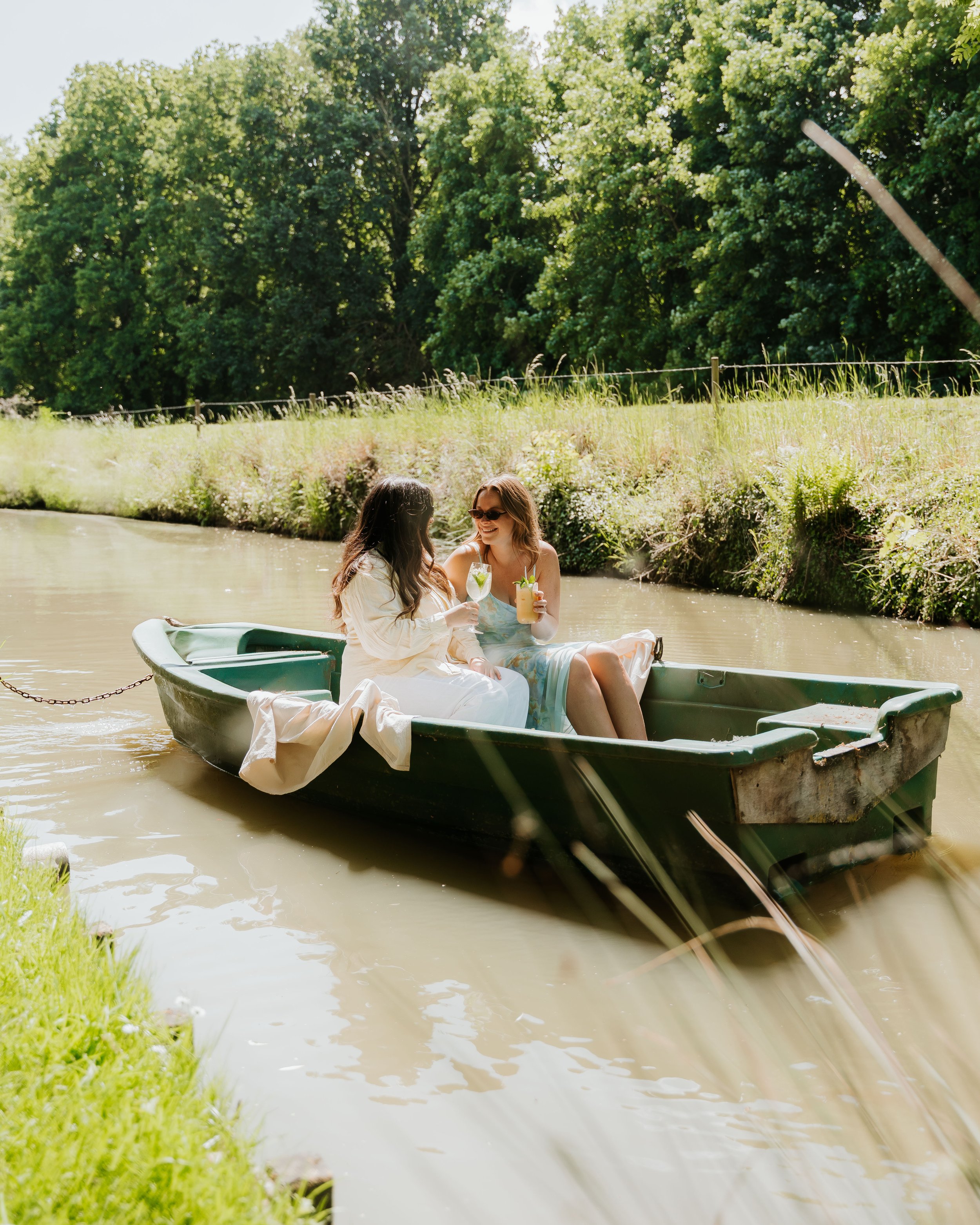 Twee vrouwen genieten in een boot op een rustige rivier, drinkend en lachend, omgeven door groene bomen en planten.