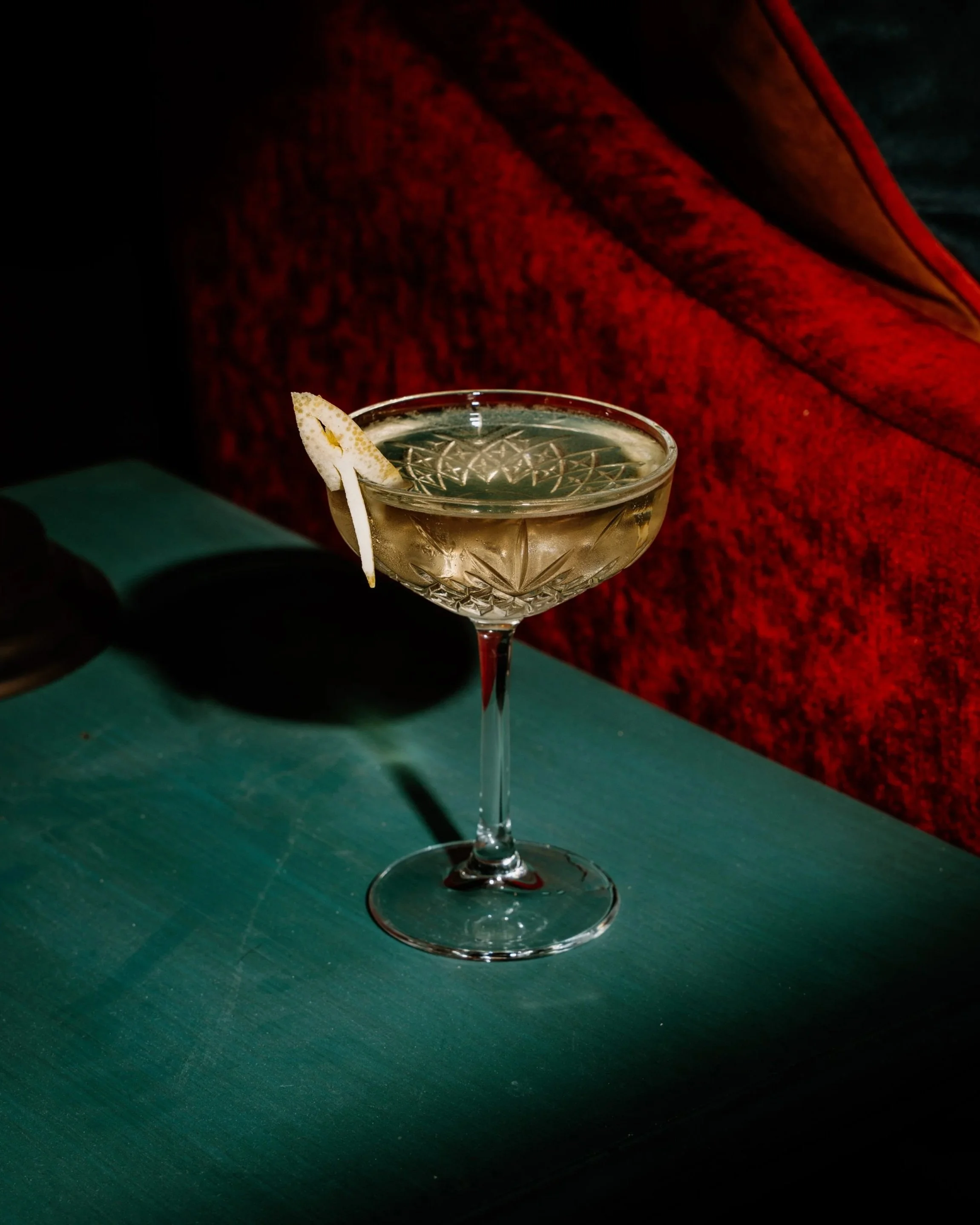 Een cocktail in een grote kristallen glas met een schelpvormige garnering op de rand, op een groen tafelpand, met een rode bekleding op de achtergrond.