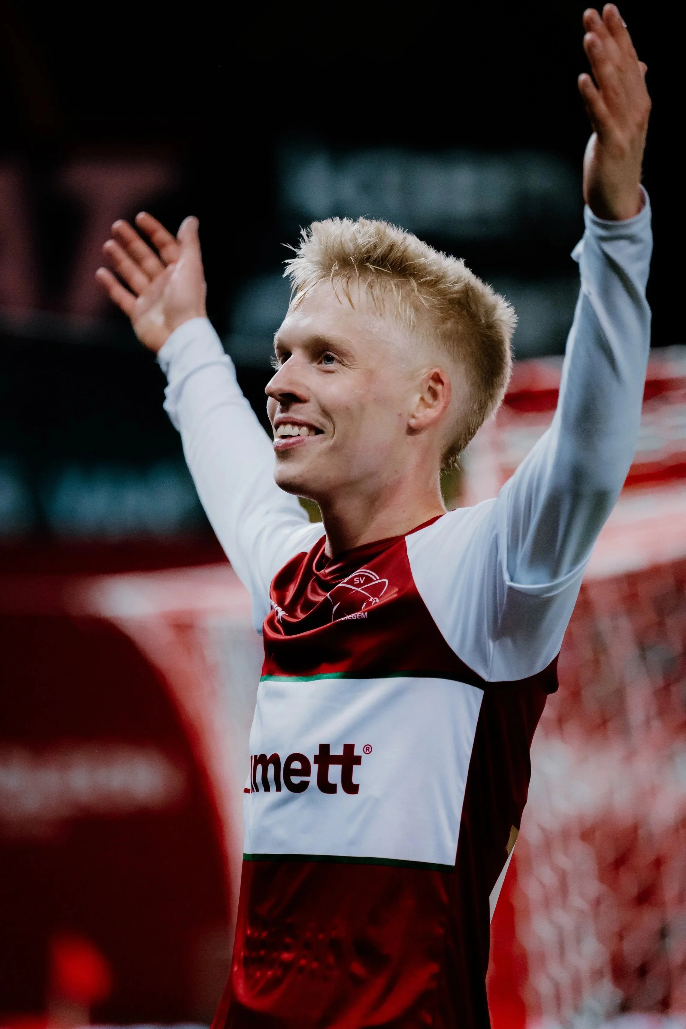 Een man in een rood-wit sportoutfit viert met opgetrokken armen op een sportveld.