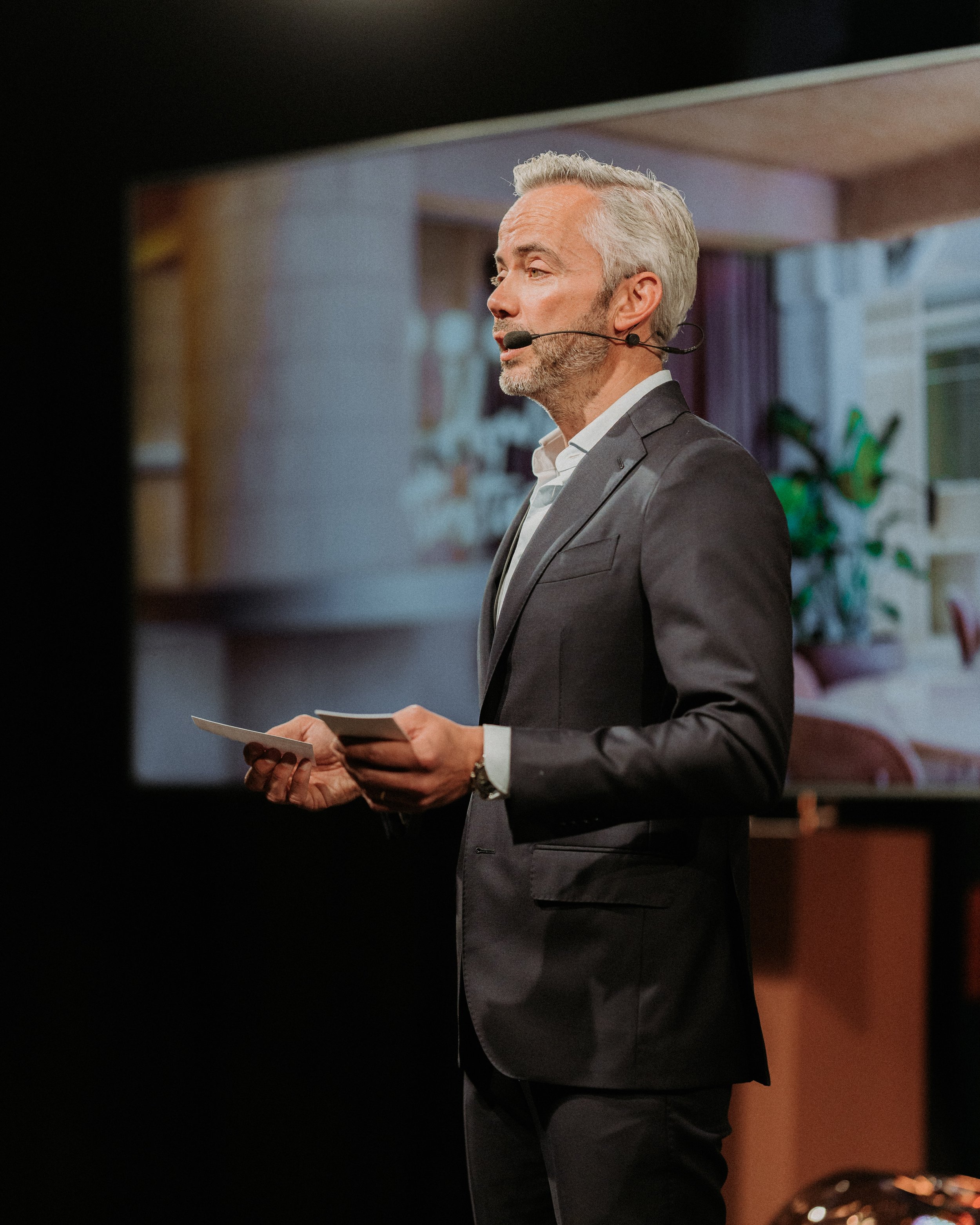 Man in pak met microfoon en notitieboekje, presentatieluidspreker, in een modern interieur, mogelijk tijdens een conferentie of presentatie.