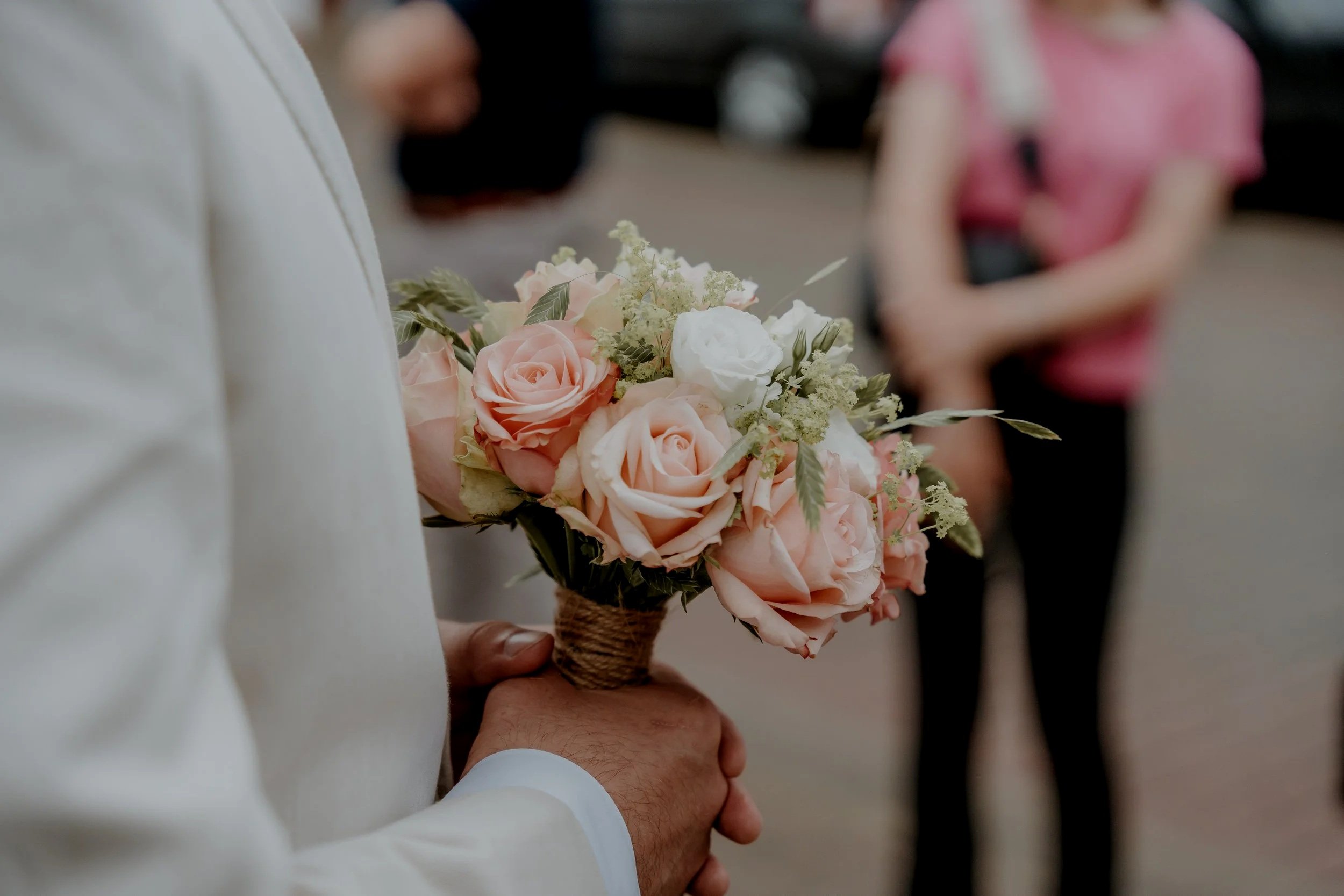 Een persoon houdt een boeket met lichtroze en witte rozen, afgebeeld van dichtbij, met andere mensen op de achtergrond in een indoorsetting.