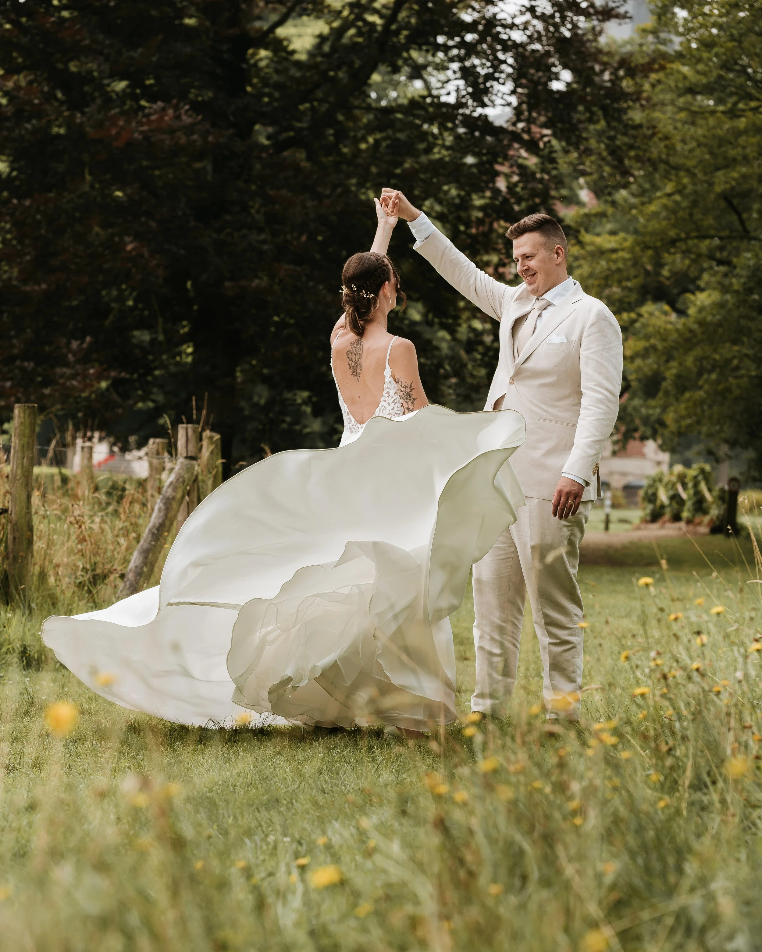 Twee mensen dansen in een tuin, een man in een witte broek en jas, een vrouw met een witte trouwjurk met open rug en fijne details, omgeven door gras en bloemen, met bomen op de achtergrond.