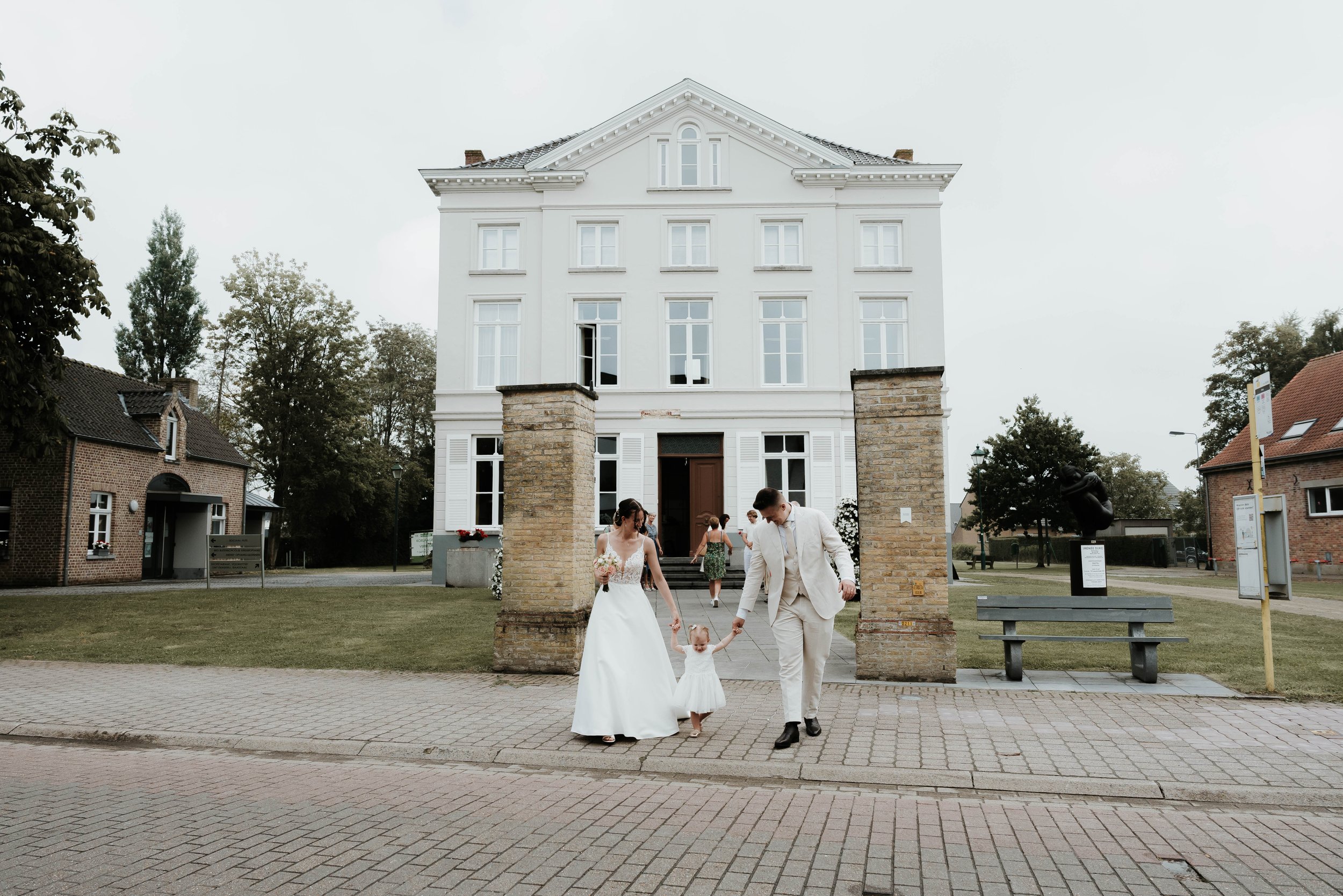 Een bruid en bruidegom lopen samen met een klein meisje, mogelijk hun dochter, op straat voor een wit gebouw, tijdens een huwelijk. De bruid draagt een witte jurk, de bruidegom een licht pak, en ze houden elkaars handen vast.