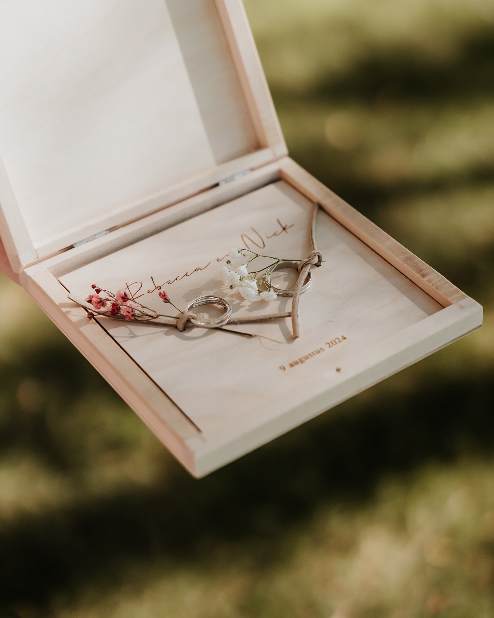 Twee trouwringen met bloemen, gelegen op een houten hart, in een open houten kist met de naam 'Rebacca en Nilt' en de datum '9 augustus 2024' erop.