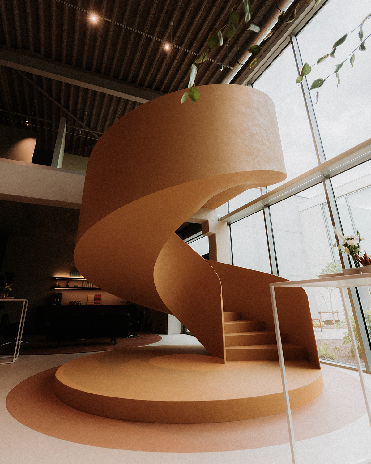Een interieur met een grote, spiraalvormige trap van bruin kartonachtig materiaal, in een modern gebouw met grote ramen en natuurlijke lichtinval.