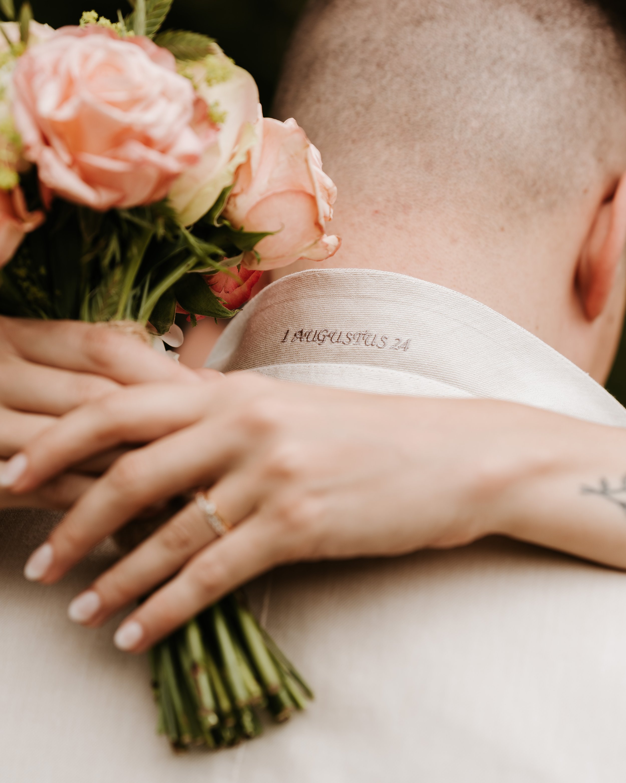 Een persoon die een bos roze rozen vasthoudt, met een ring op haar vinger. De persoon heeft een tatoeage op haar onderarm en draagt een witte kledingstuk met een inscriptie. De achtergrond is vaag.