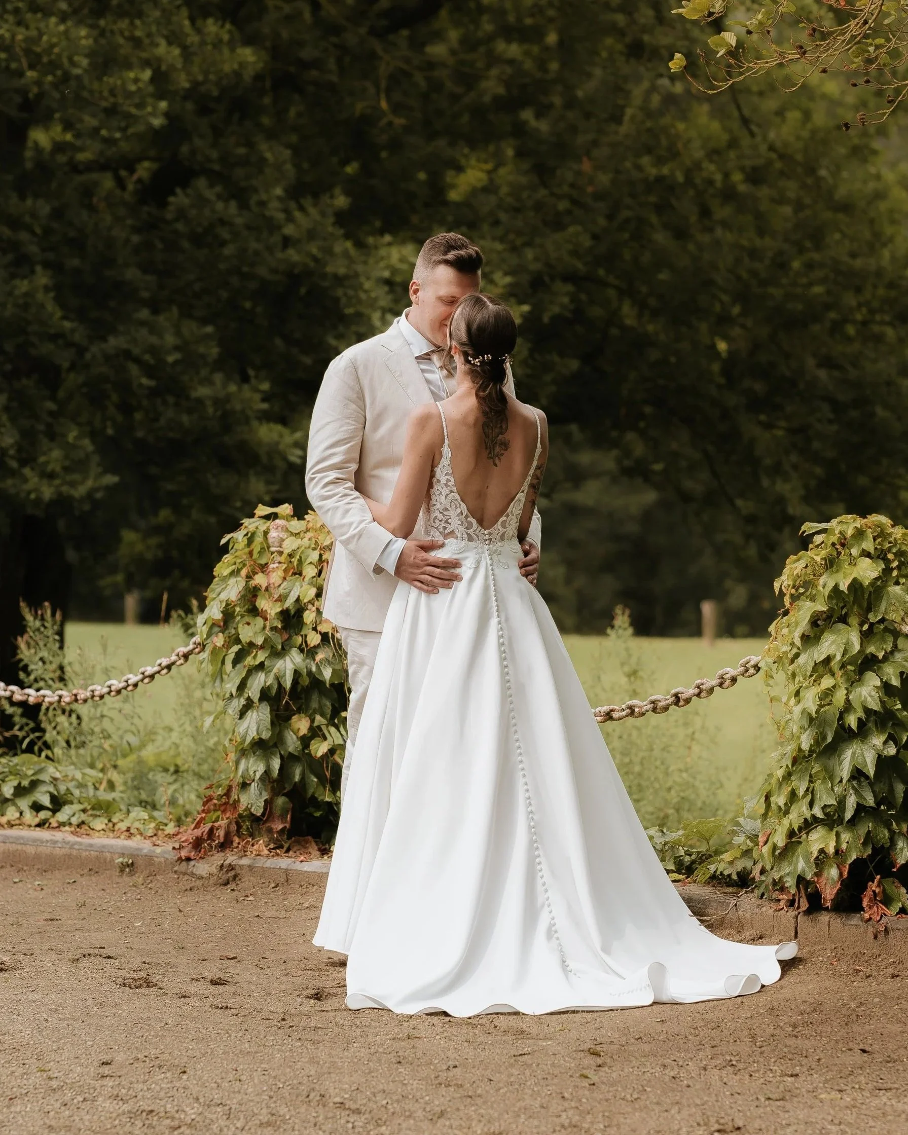 Een bruid en bruidegom staan samen in een park, omringd door groen, terwijl ze naar elkaar kijken en zich vasthouden.
