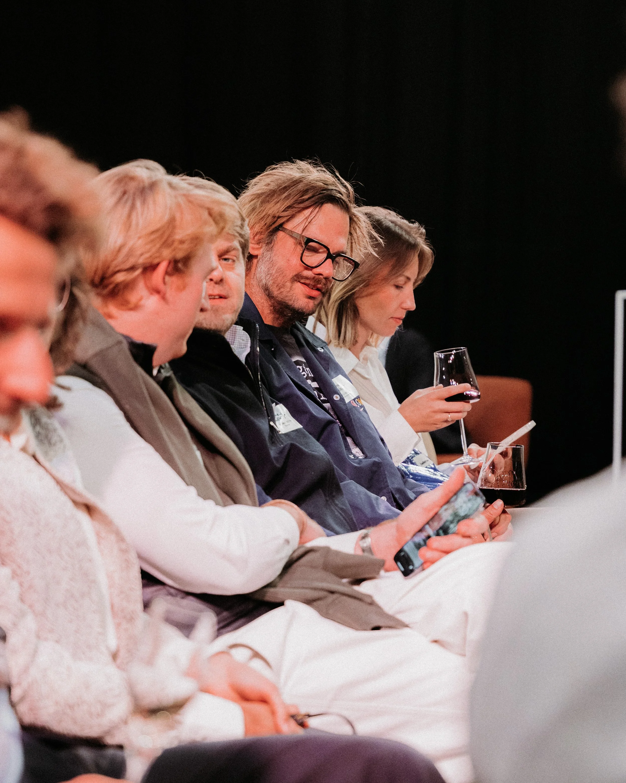 Vrouwen en mannen zitten in een rij, sommigen kijken op hun telefoons, anderen drinken wijn, op een donkere achtergrond.