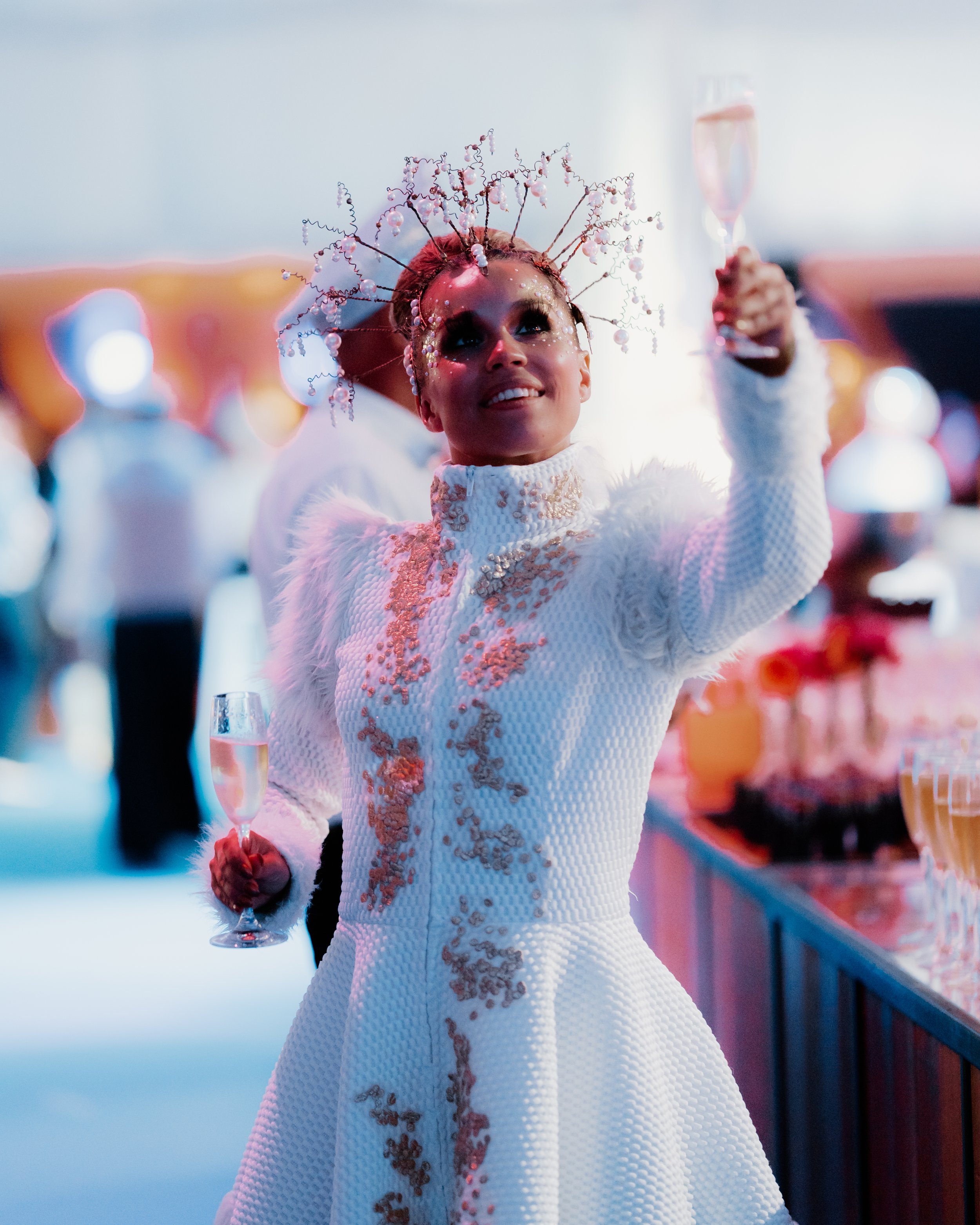 Vrouw in een witte feestjurk met roze en gouden details, met een sierlijk hoofddeksel, houdt een champagneglas en lijkt te genieten op een feest met kleurrijke verlichting.