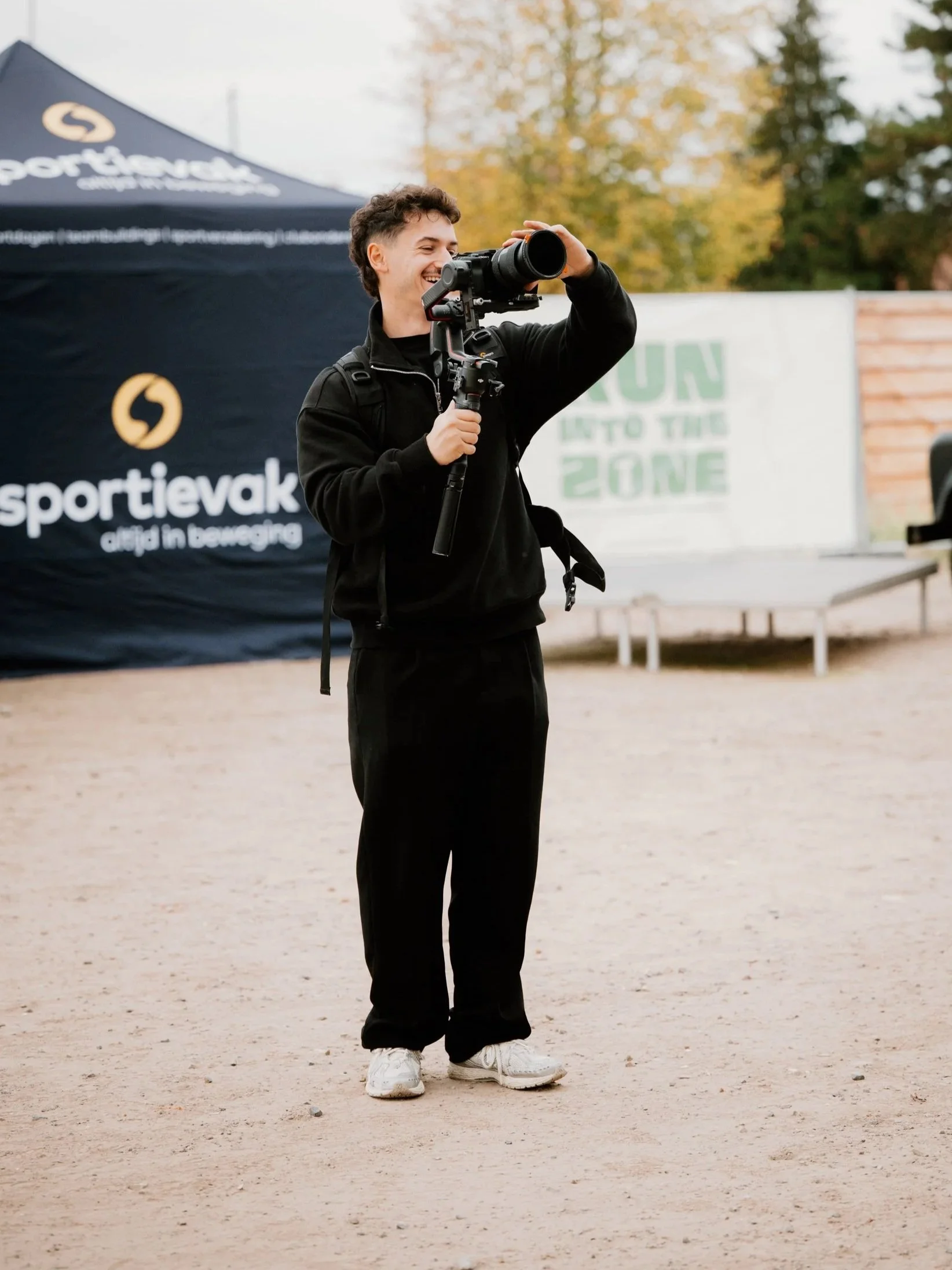 Een jongeman in een zwart trainingspak met een rugzak fotografeert met een grote telelens op een statief op een open terrein met een blauwe tent en een muur met tekst.